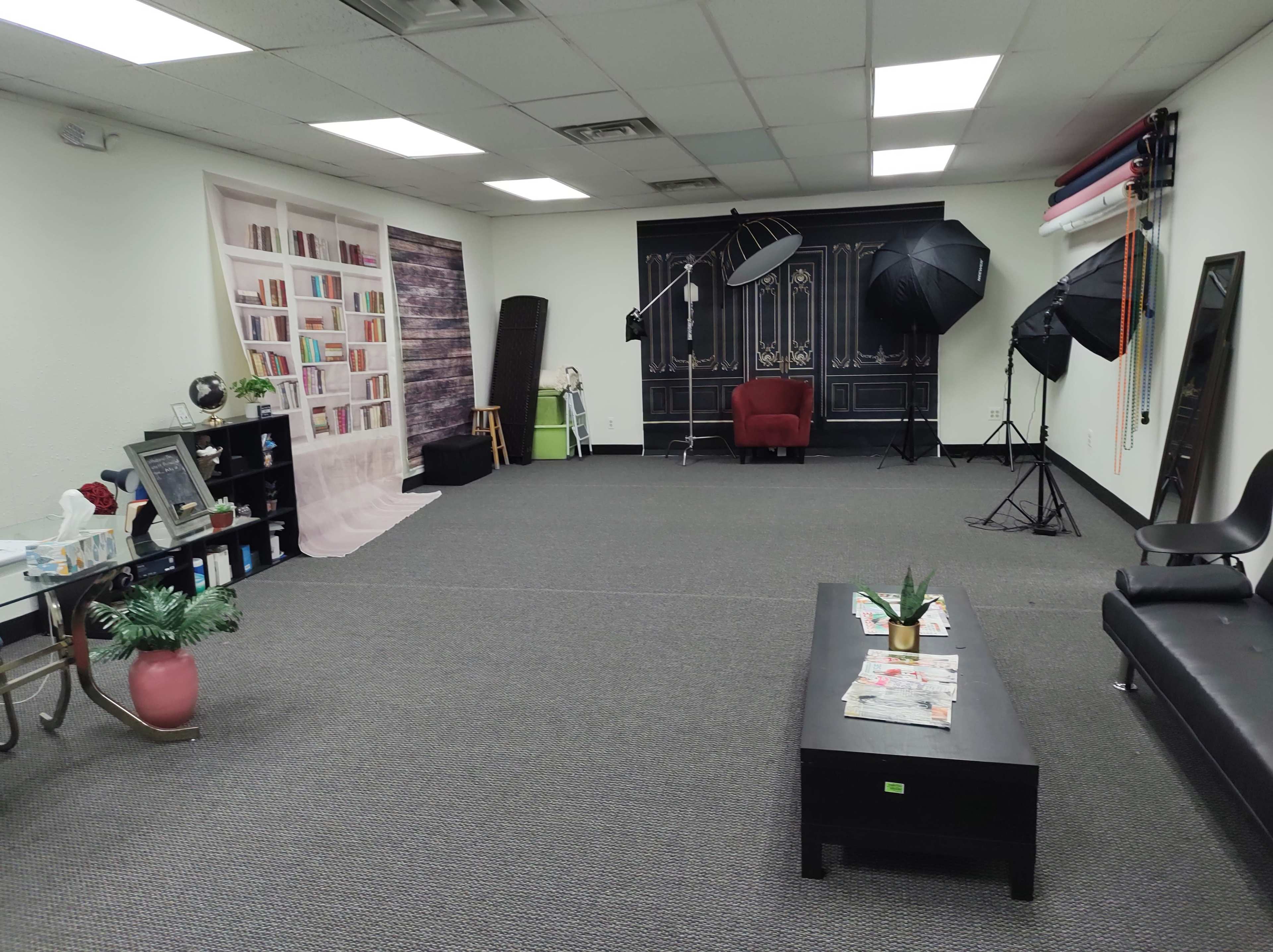 A spacious studio with a bookcase backdrop, a red armchair, photography lighting equipment, and a low table with magazines.