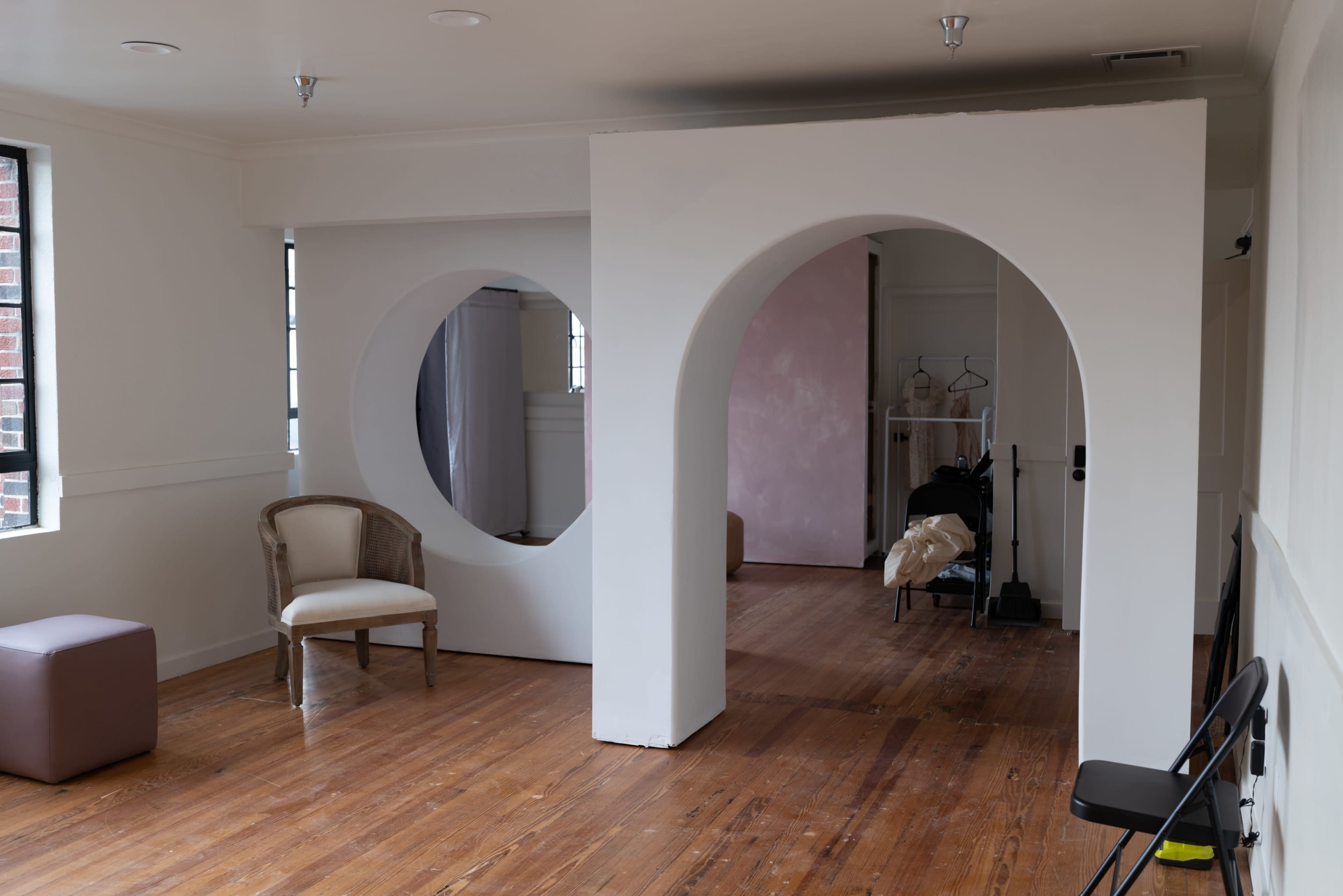 The room features two large circular archways in a white wall, with wooden floors and minimal furniture, including a chair and an ottoman.