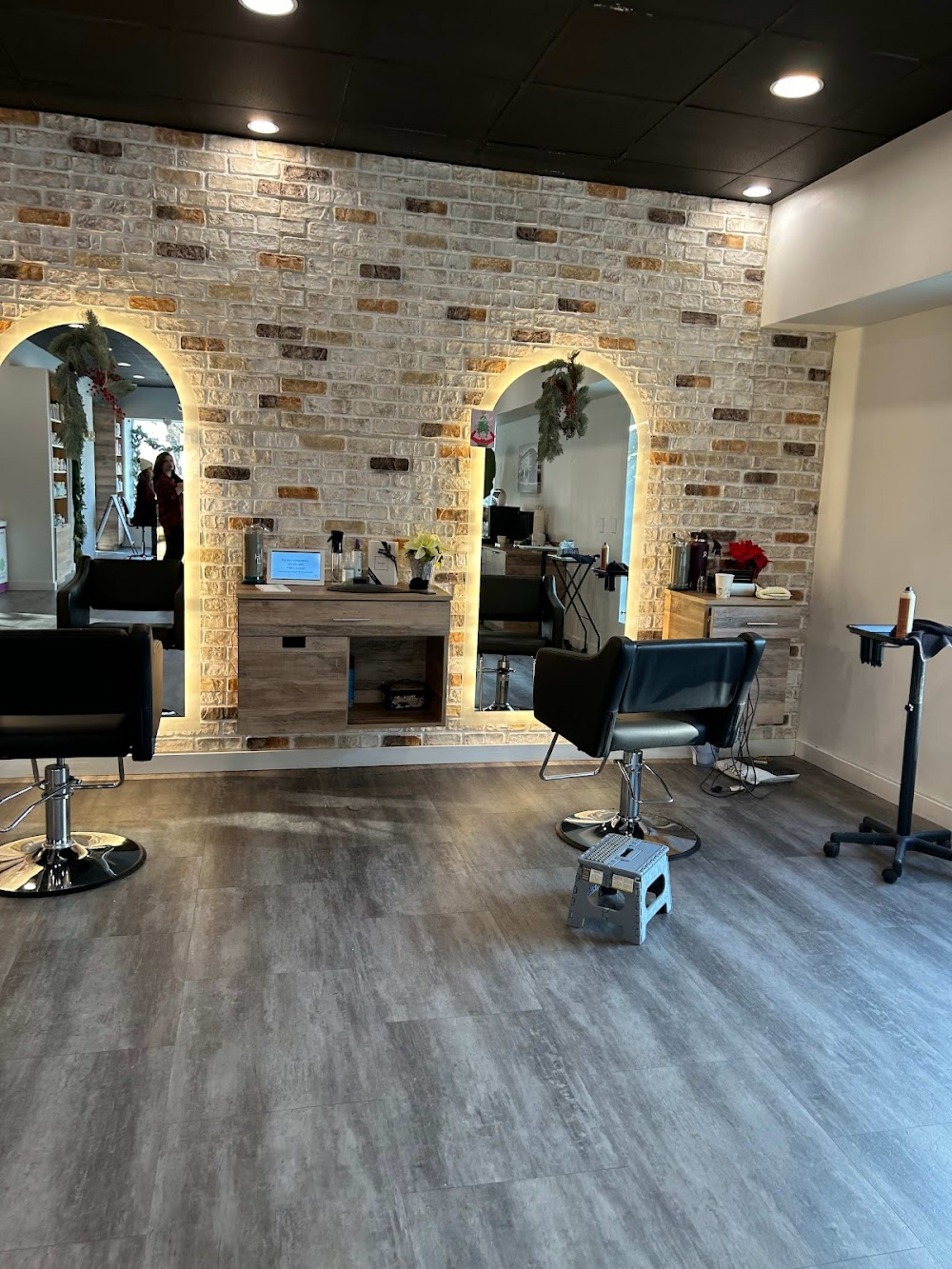 The image shows a modern salon interior with two styling stations illuminated by lighted arches, featuring black salon chairs and a wooden storage unit against a textured brick wall.