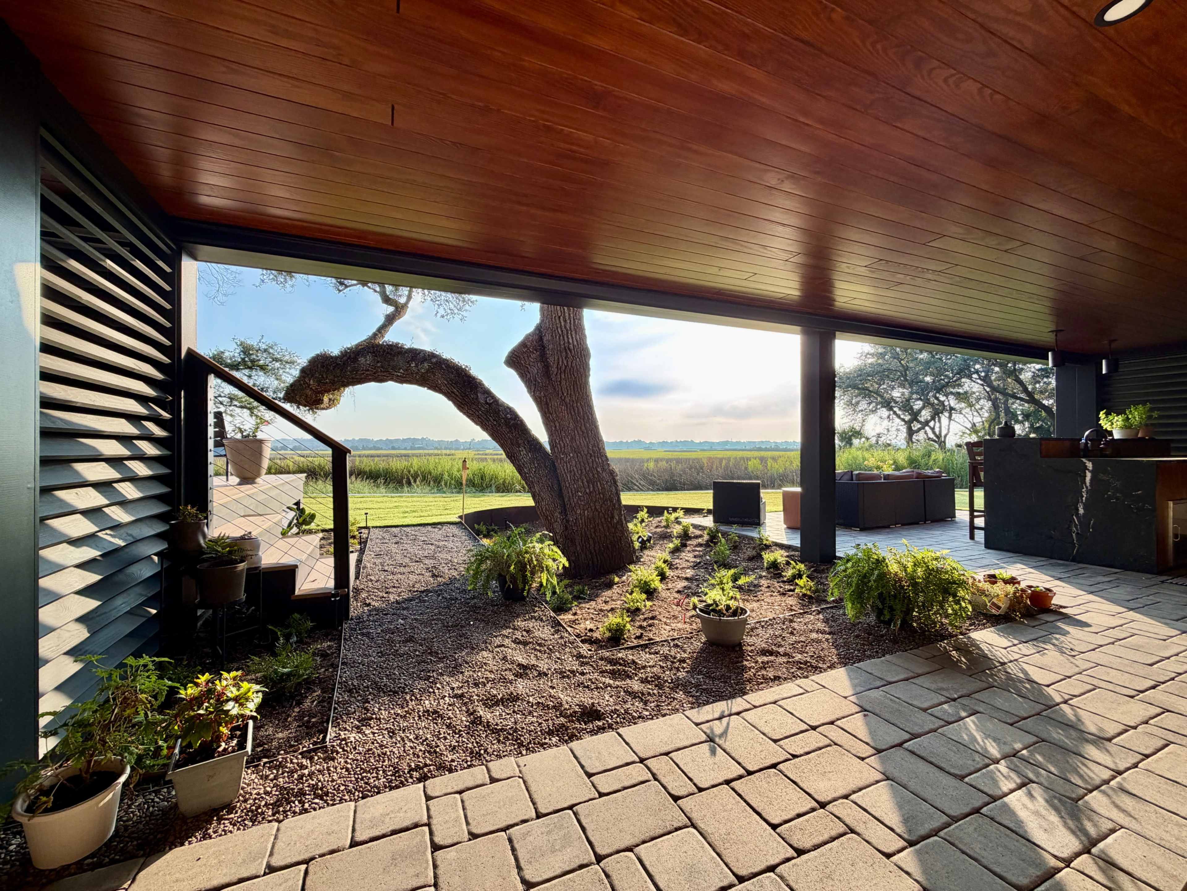 The image shows a modern outdoor living space featuring a wooden ceiling, planter beds, and a scenic view of a grassy field and trees.
