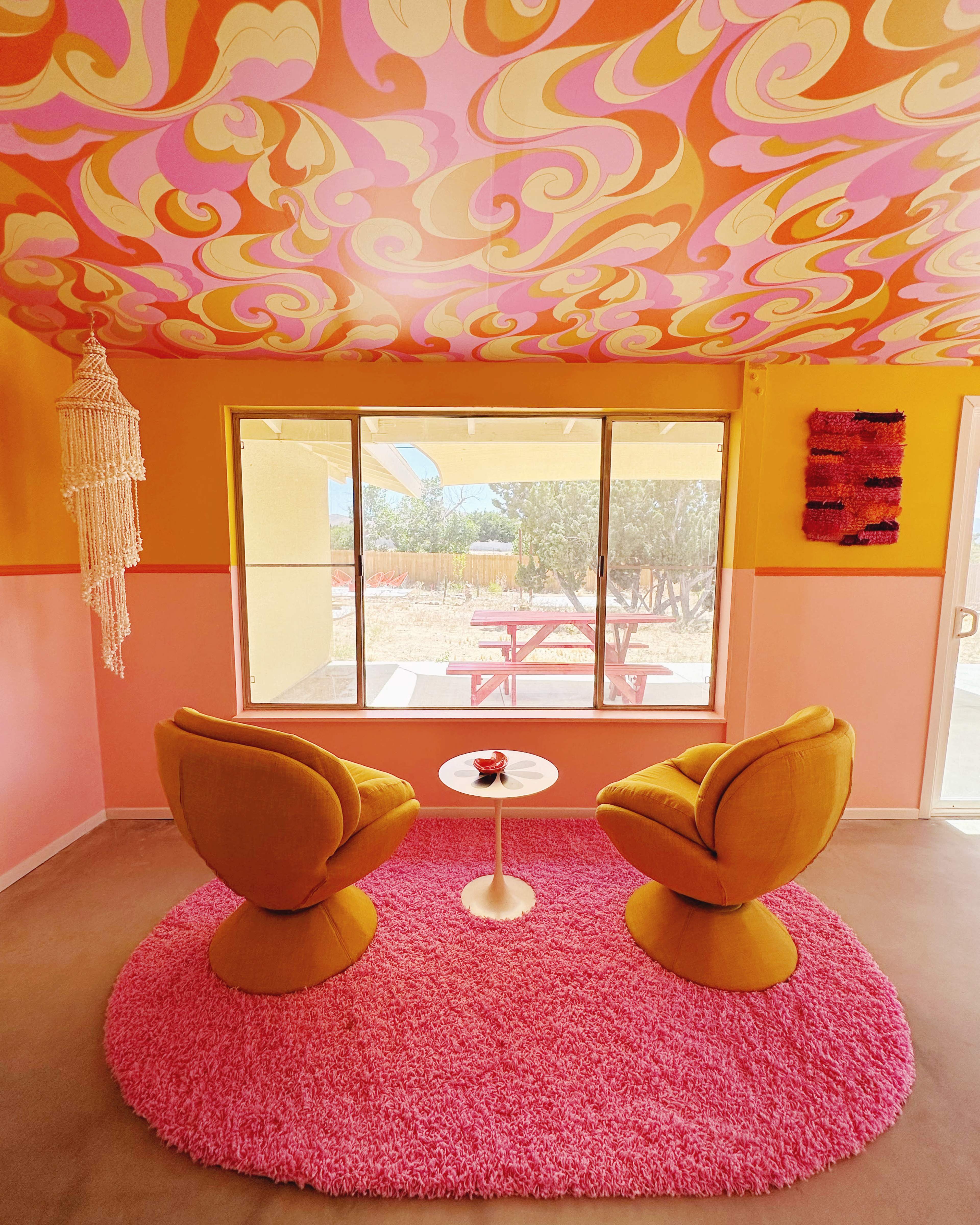The scene features two yellow chairs and a small white table on a pink rug, set against a backdrop of a brightly patterned ceiling and a large window showcasing an outdoor picnic area.