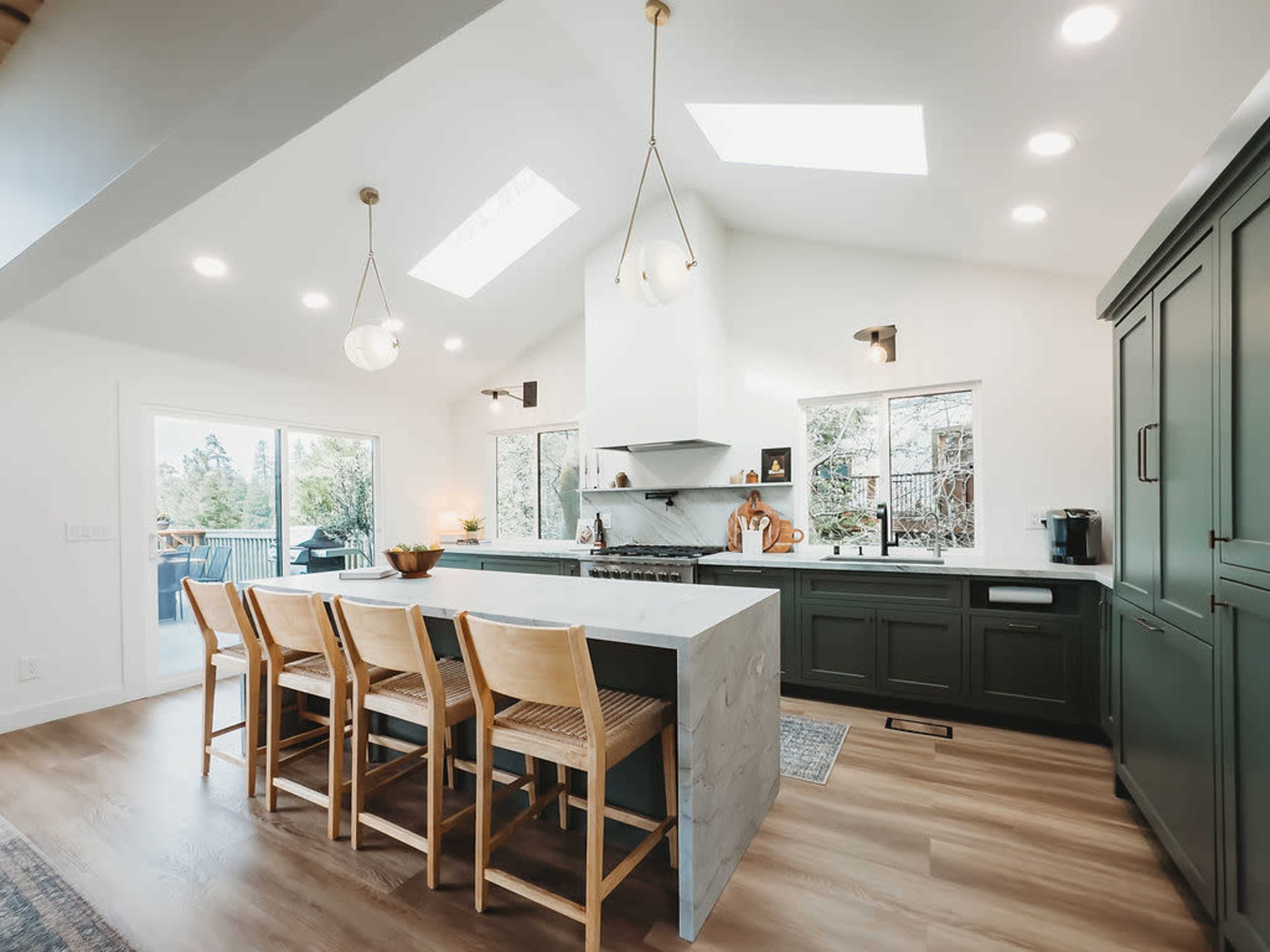 A modern kitchen features a central island with seating, large windows for natural light, and minimalist cabinetry in green and white.