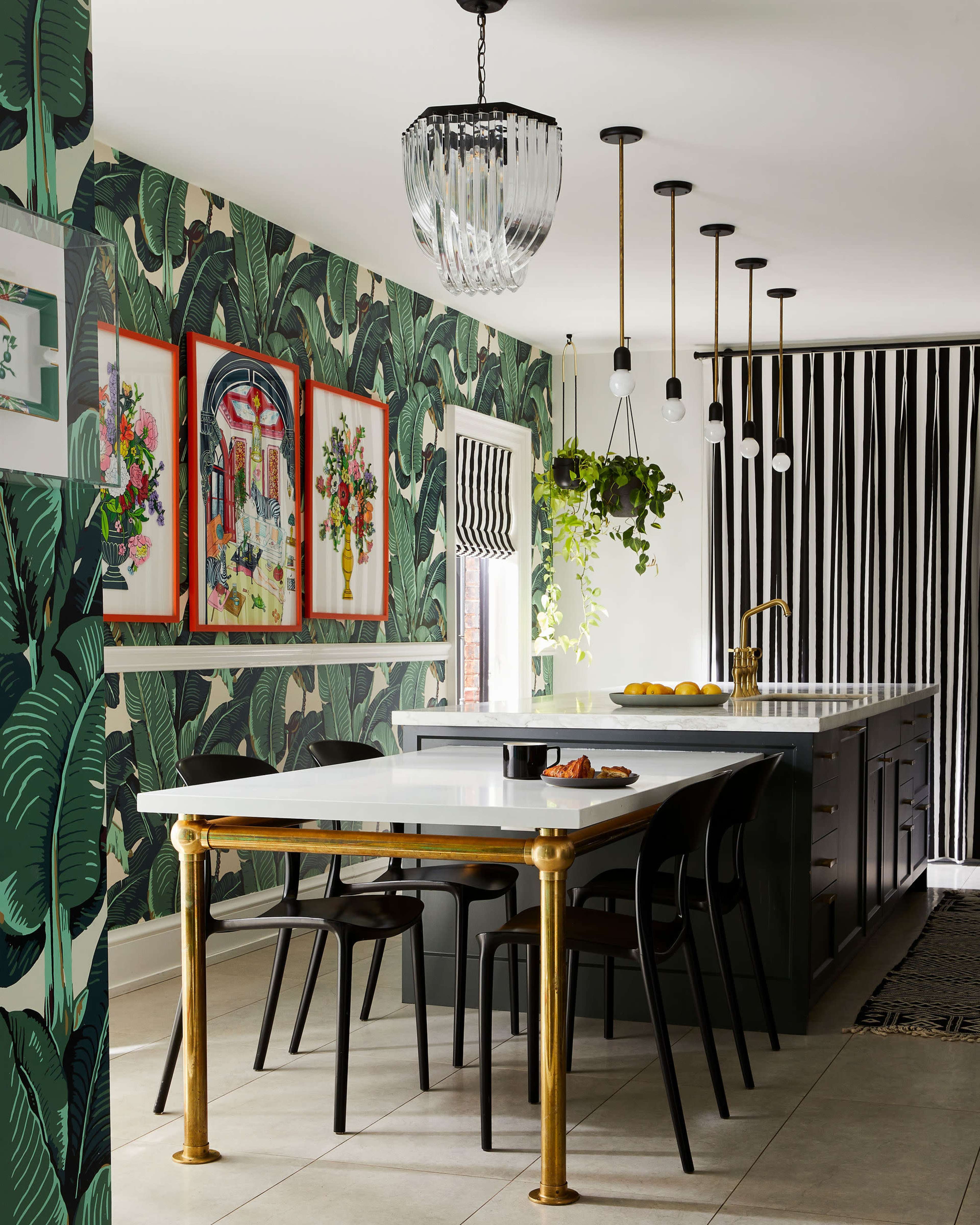The kitchen features a long island with a white countertop, black cabinetry, and bold green foliage wallpaper, accented by decorative artwork and a modern chandelier.