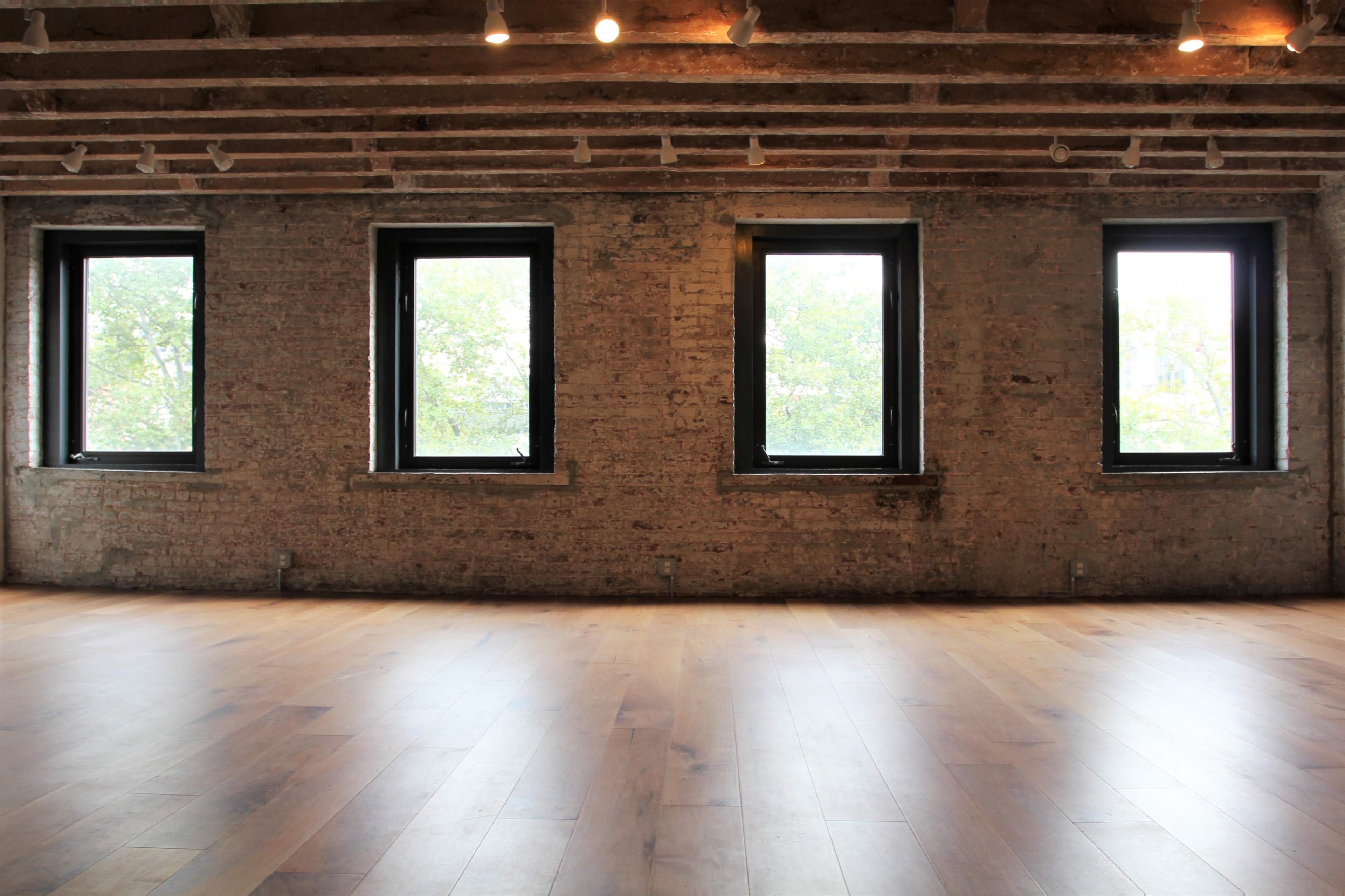 A spacious room with three large windows set in a brick wall, featuring wooden flooring and exposed ceiling beams.
