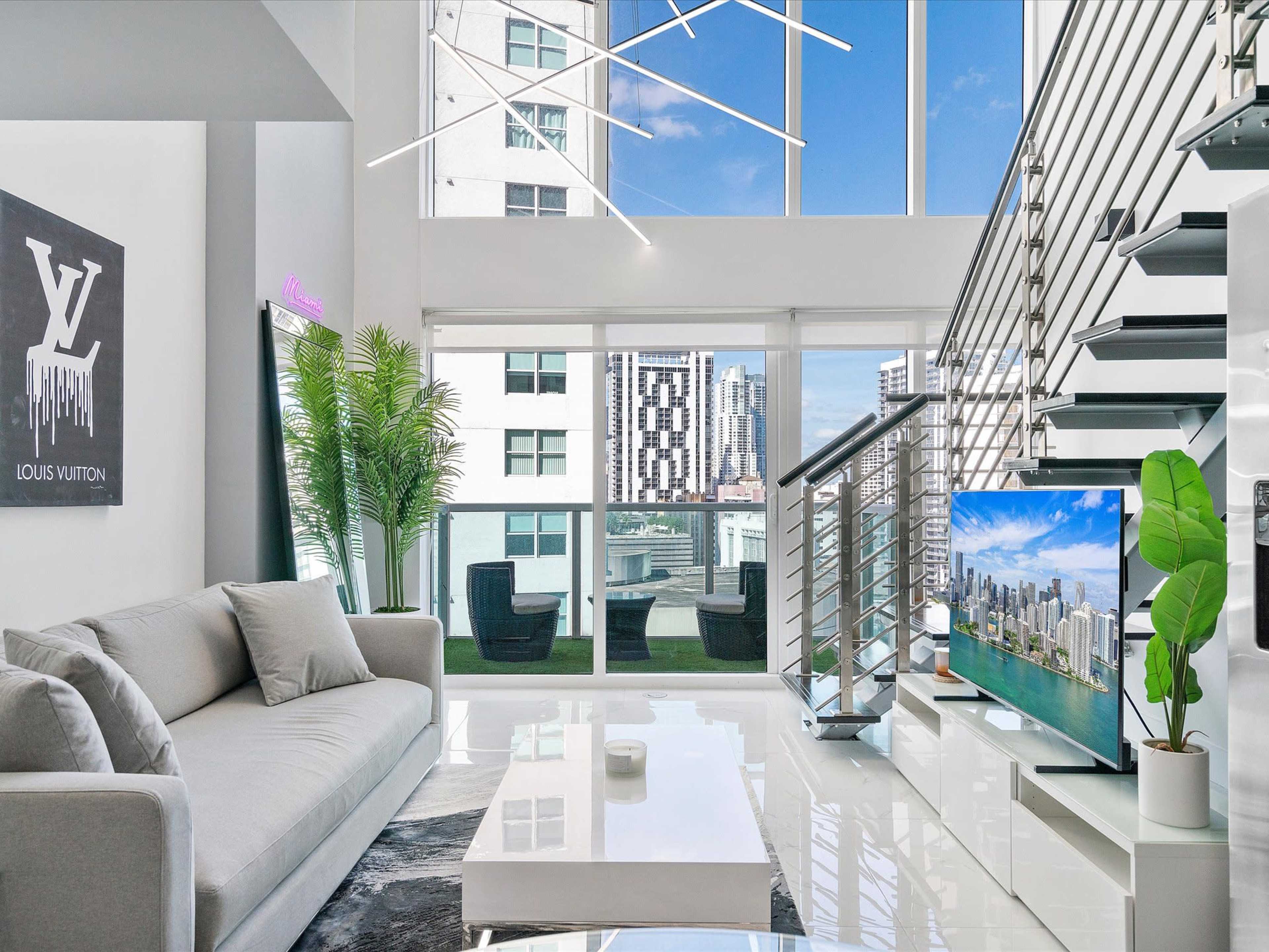 A modern living room with large windows, a sleek gray sofa, a glass coffee table, and a staircase leading to an upper level, overlooking a city skyline.