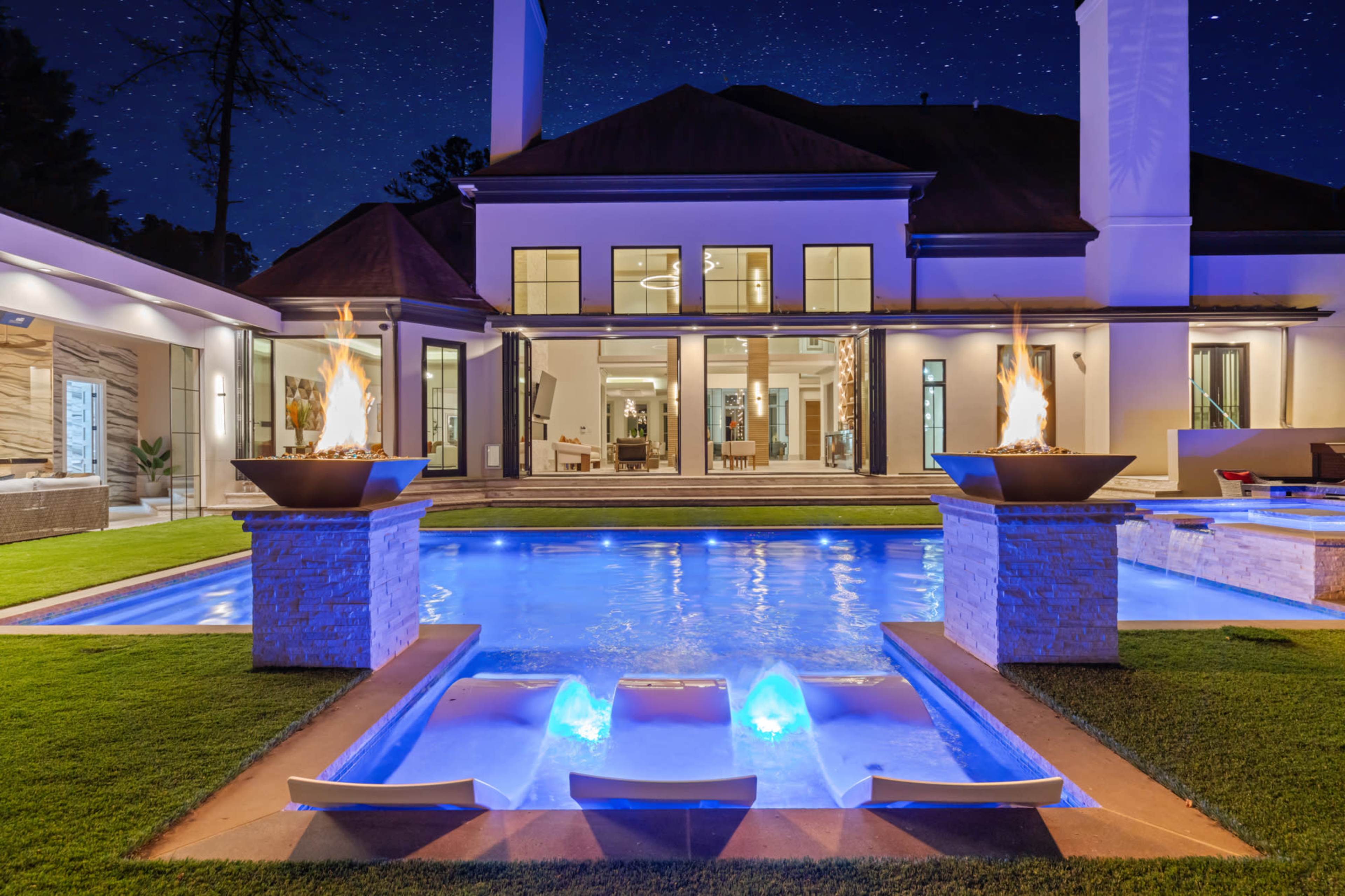 The image depicts a luxurious home with a swimming pool featuring illuminated water features, surrounded by well-manicured grass and framed by outdoor lighting amidst a starry night sky.