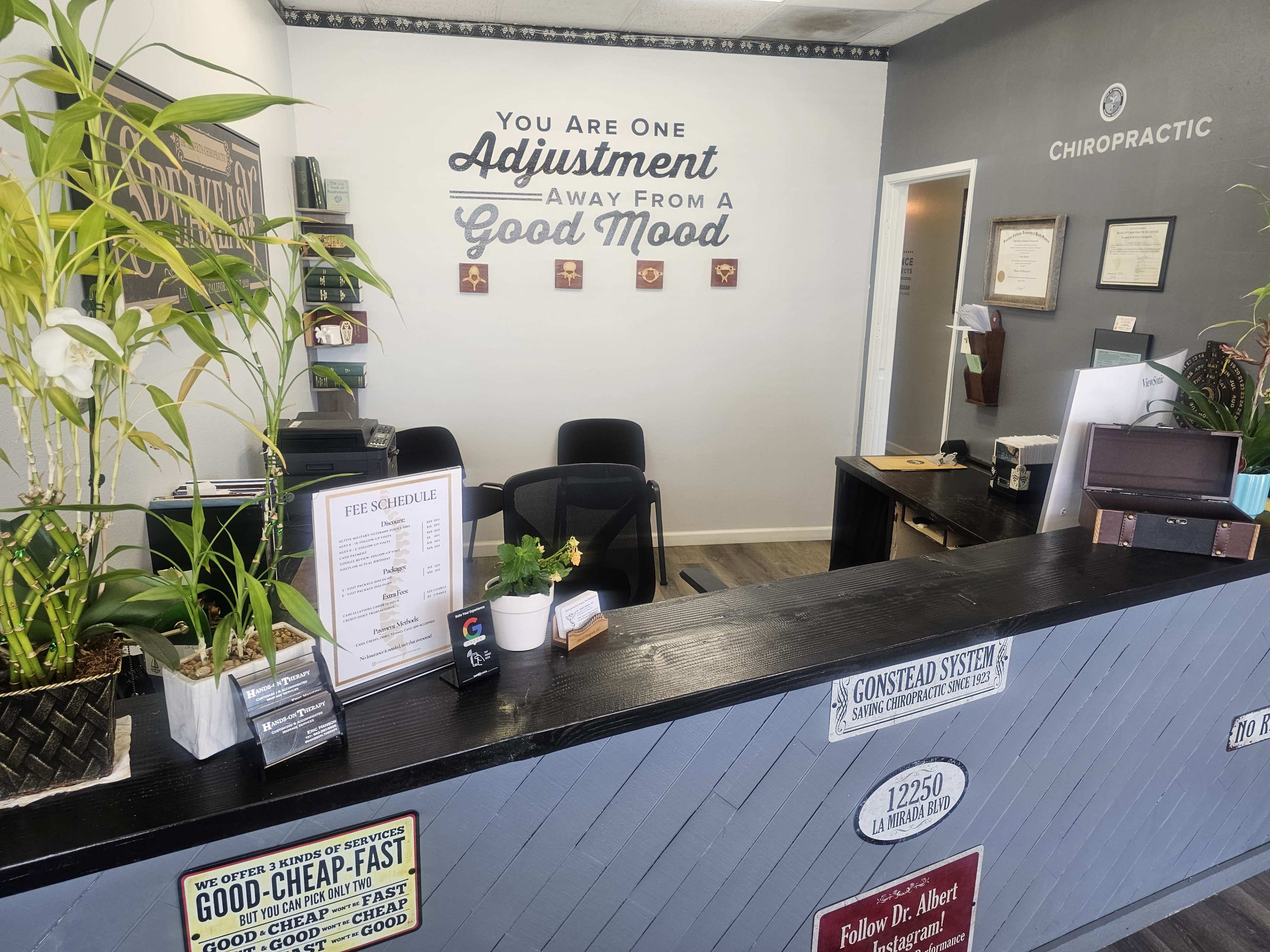 The image shows the reception area of a chiropractic office, featuring a front desk, chairs, and a motivational quote displayed on the wall.