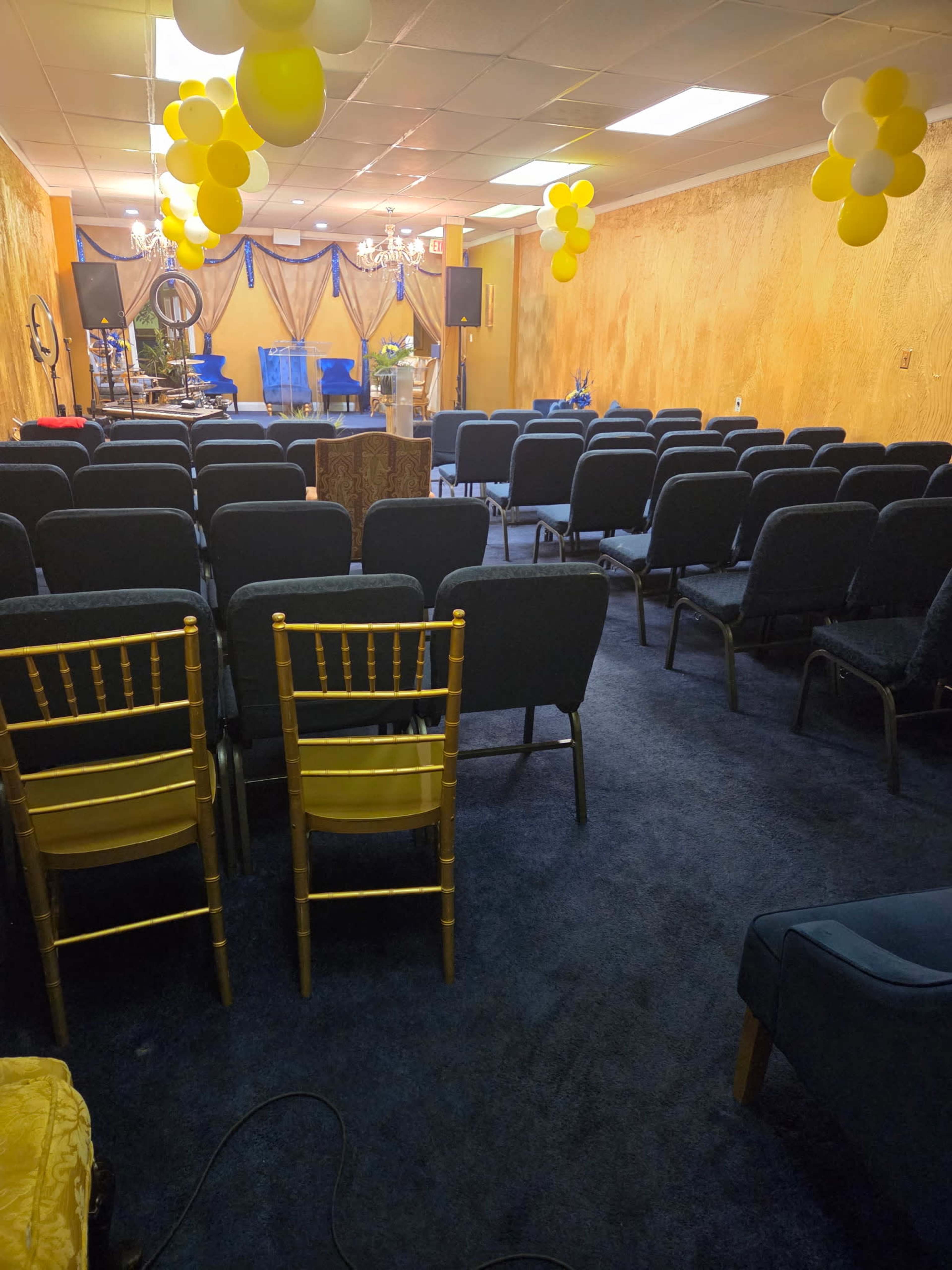 The image shows a room arranged with rows of chairs facing a stage, decorated with yellow balloons and drapery.