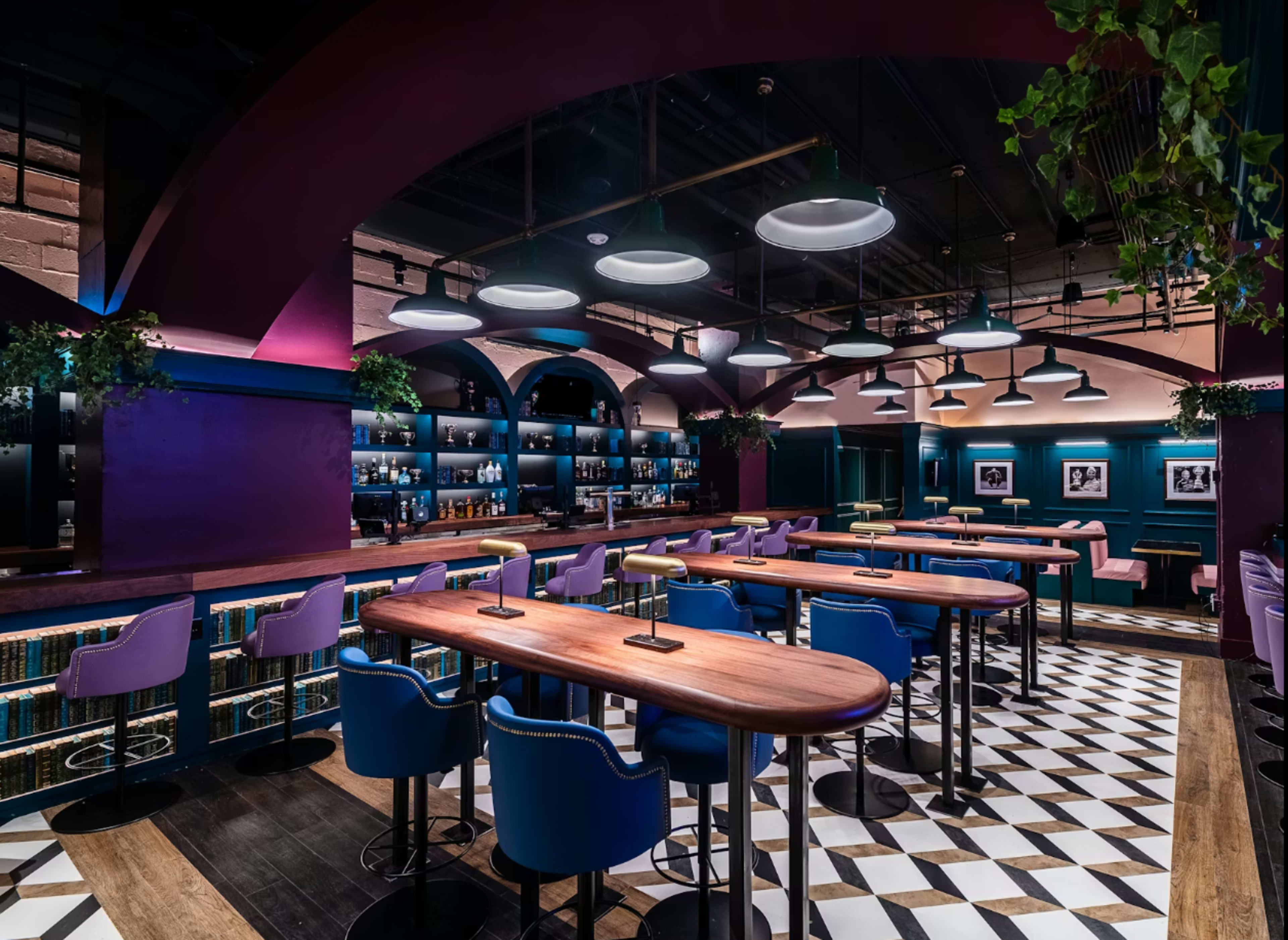 A modern bar interior features a mix of high tables and colorful seating under stylish overhead lights, with a sleek backdrop of shelves displaying bottles.
