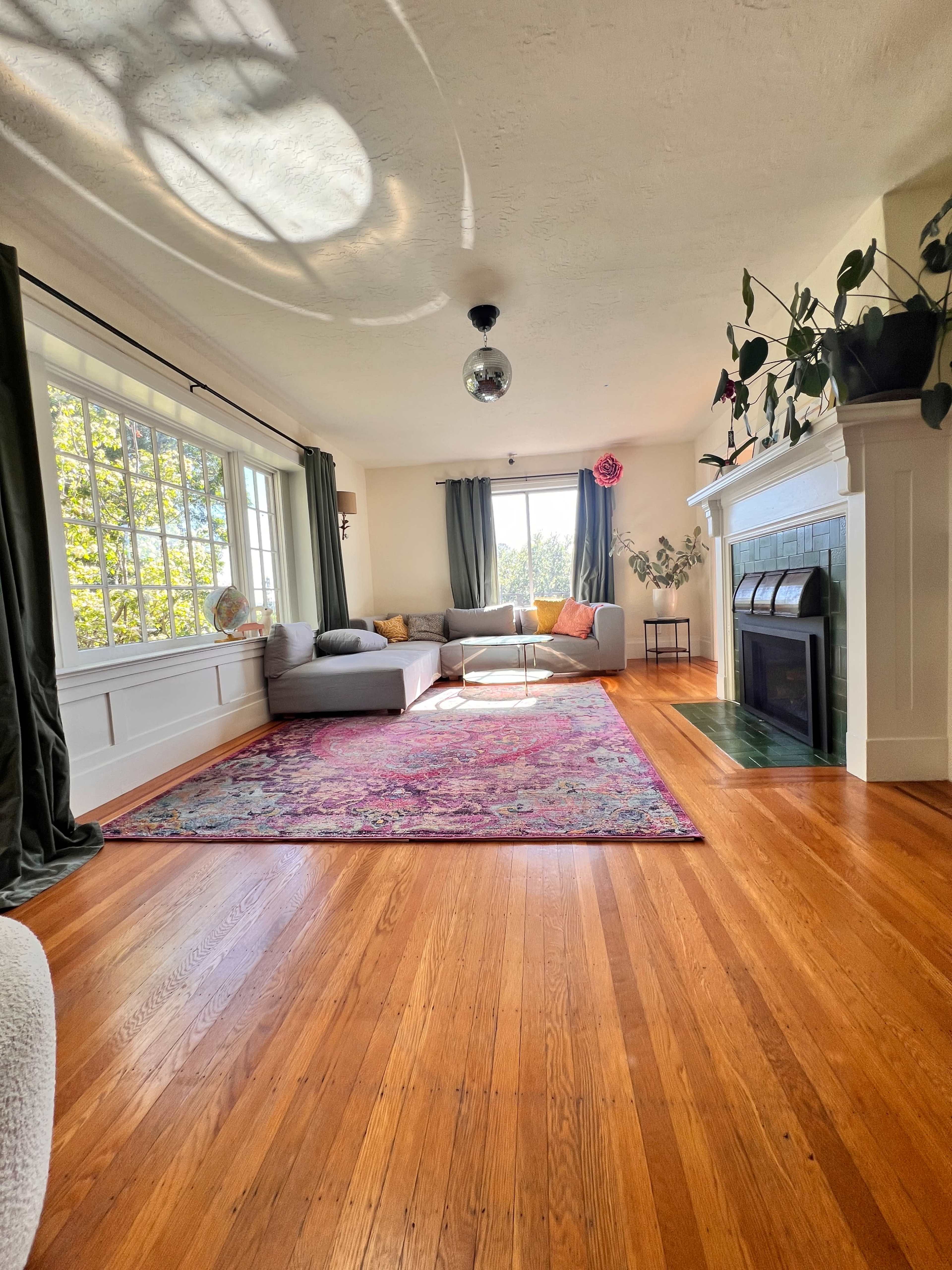 The image shows a brightly lit living room with a large window, a sectional sofa, a colorful area rug, and a fireplace.