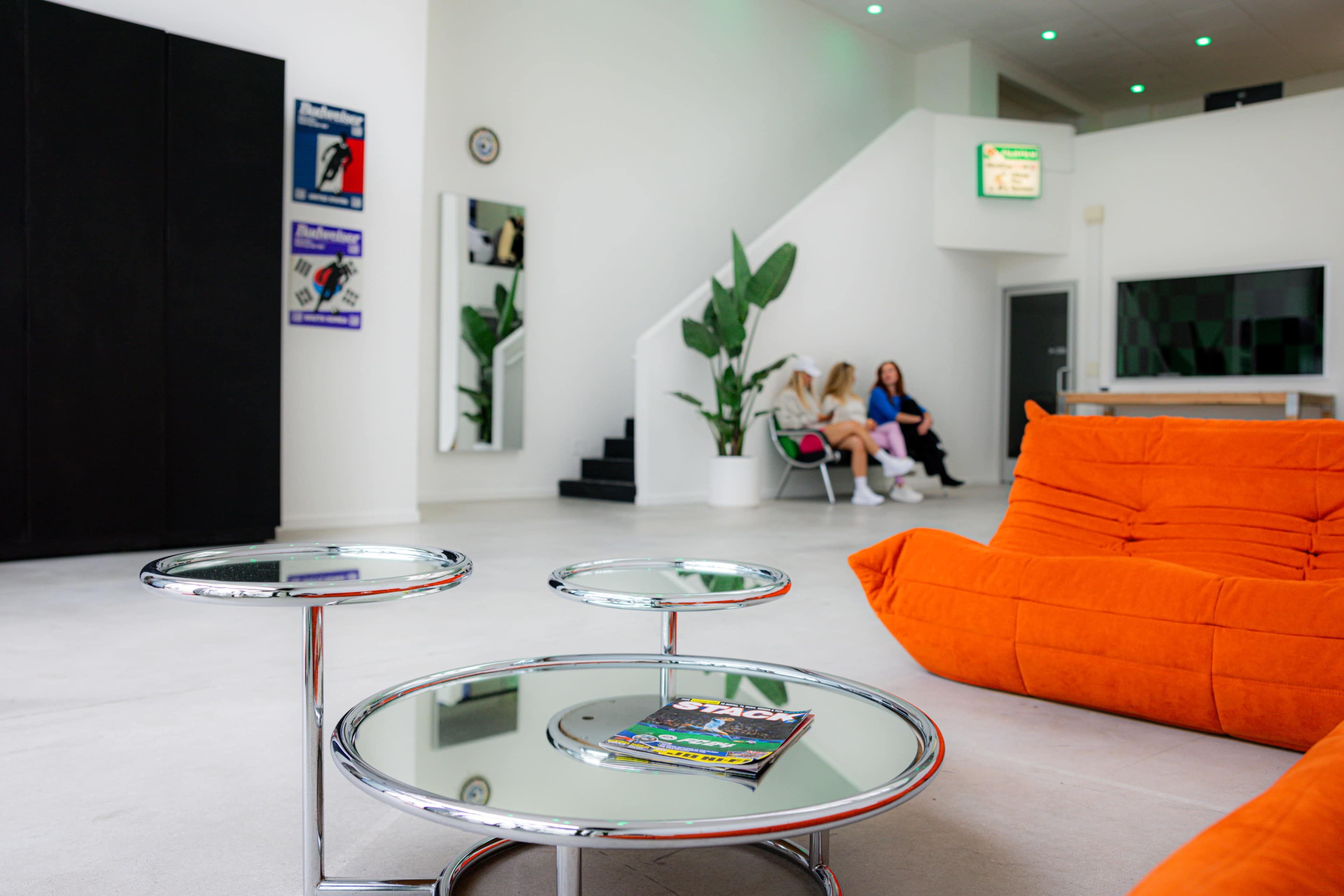 A modern interior features an orange couch and a glass coffee table, with two people seated in the background near a staircase.