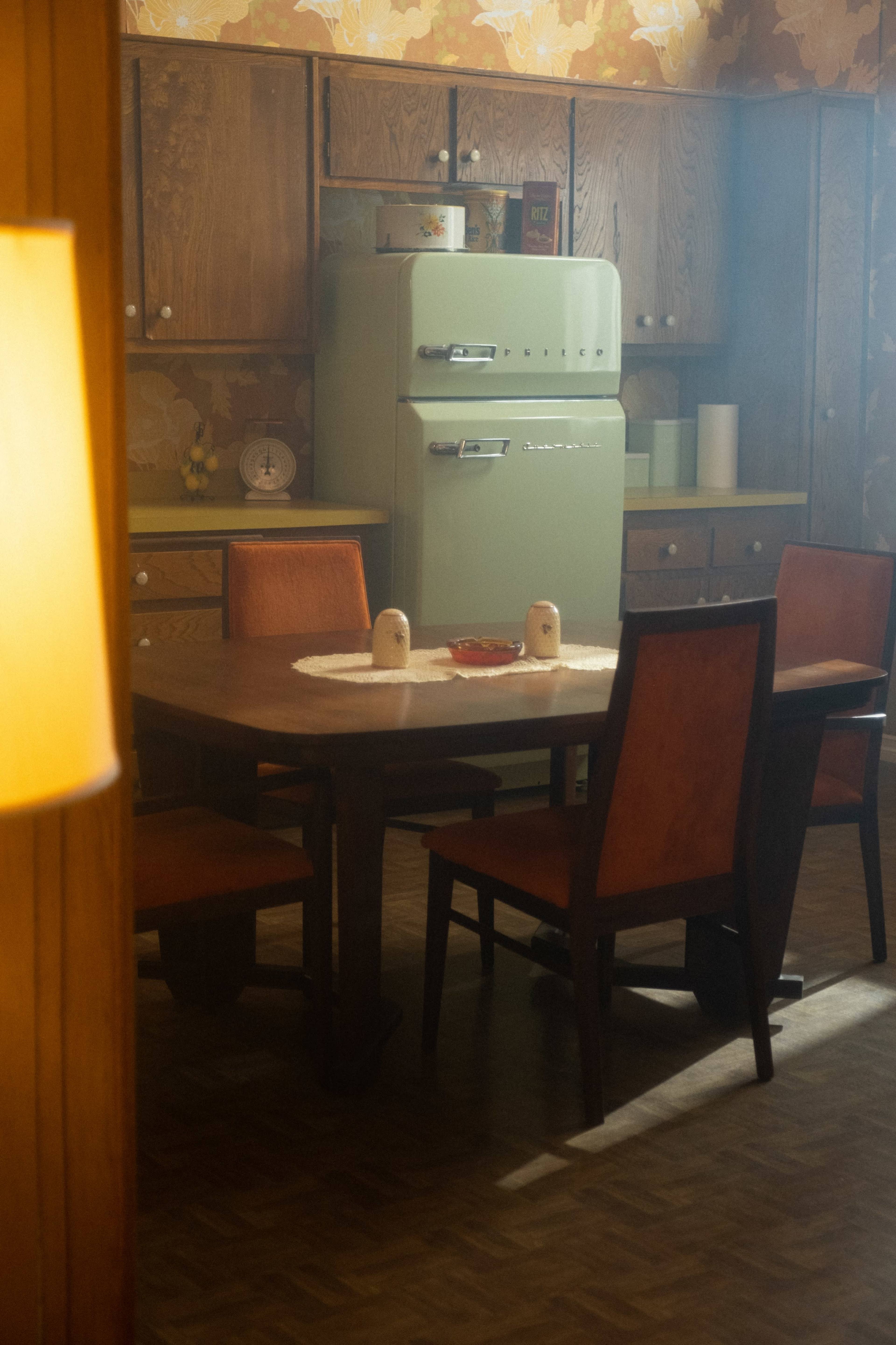 A retro kitchen features a green refrigerator, a wooden dining table with four chairs, and a floral-patterned wallpaper.