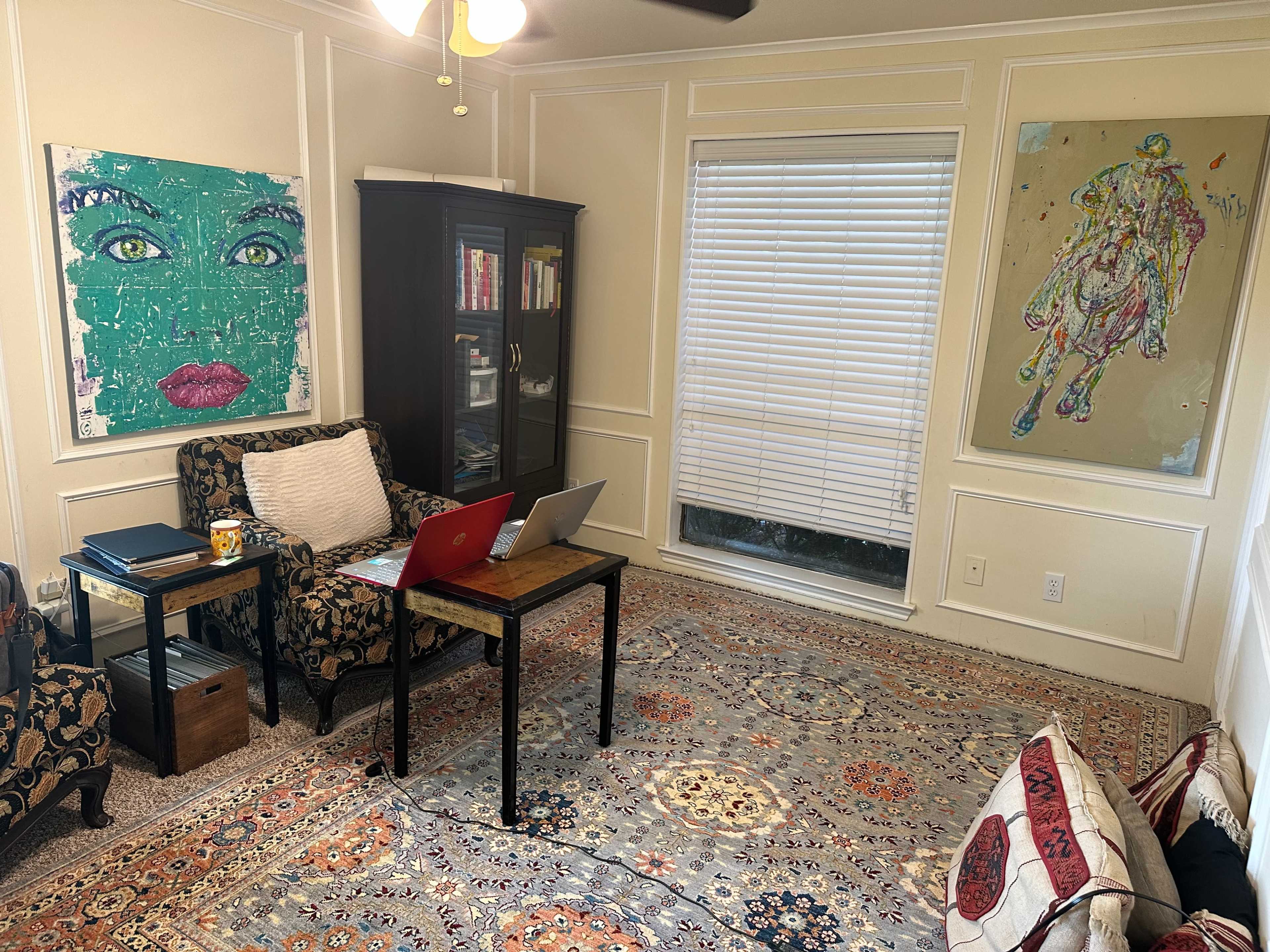 The image depicts a cozy room with a table and laptop, two patterned armchairs, and colorful artwork on the walls.