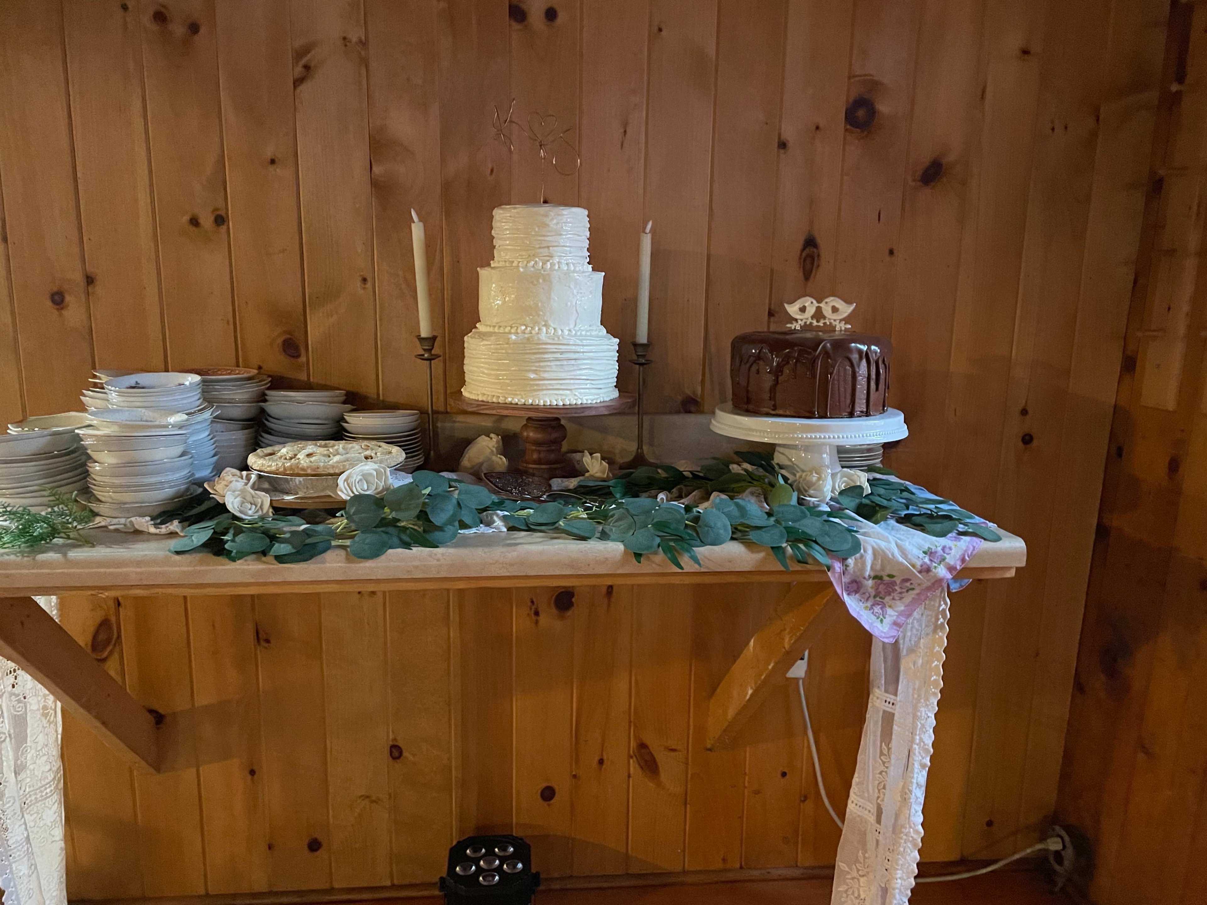 A wooden table displays two wedding cakes, a white tiered cake and a chocolate cake, surrounded by plates and decorative greenery.