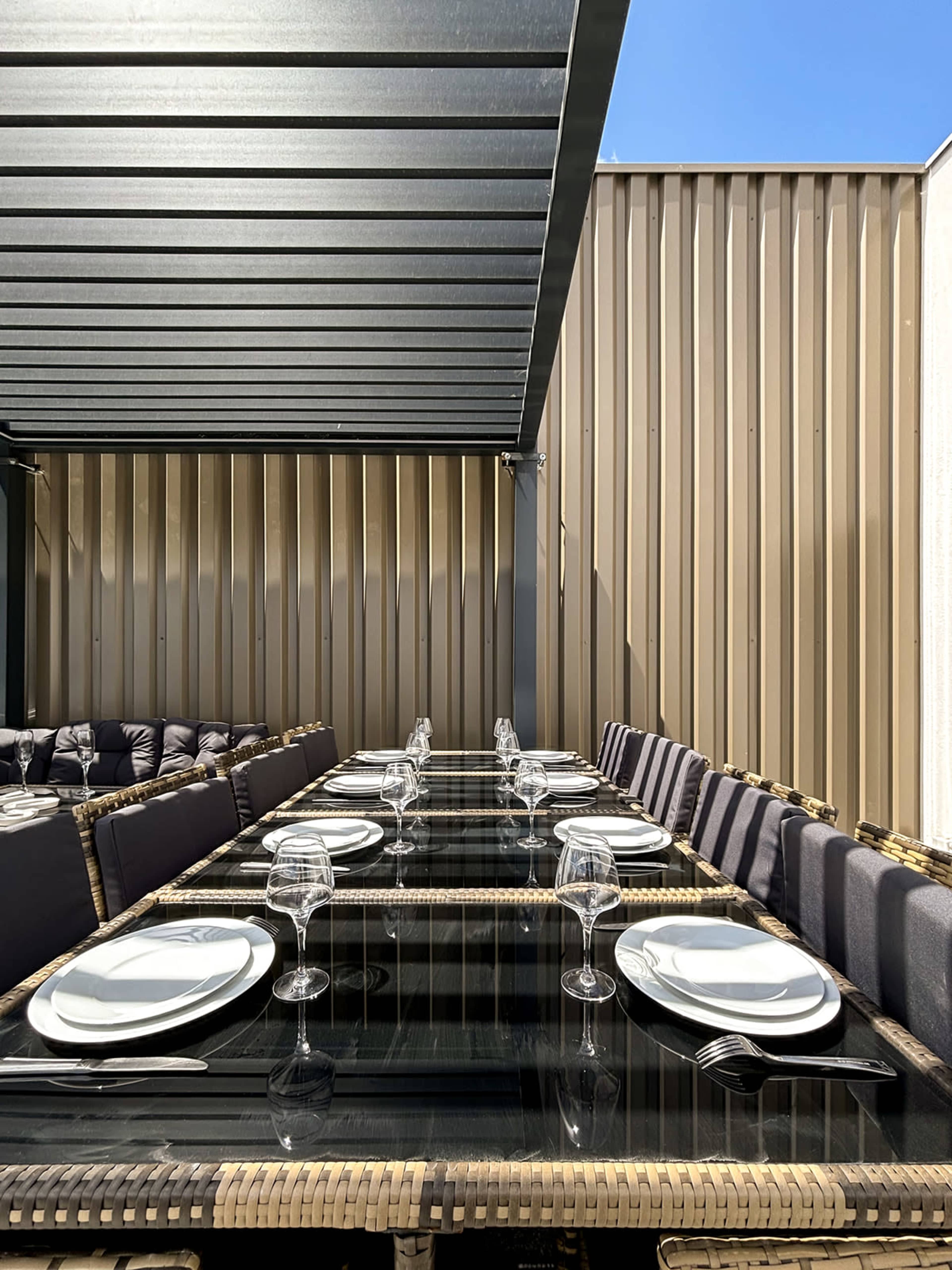 A long dining table is set under a covered patio, featuring black glass plates, white dishes, and glassware, with a backdrop of a corrugated metal wall.