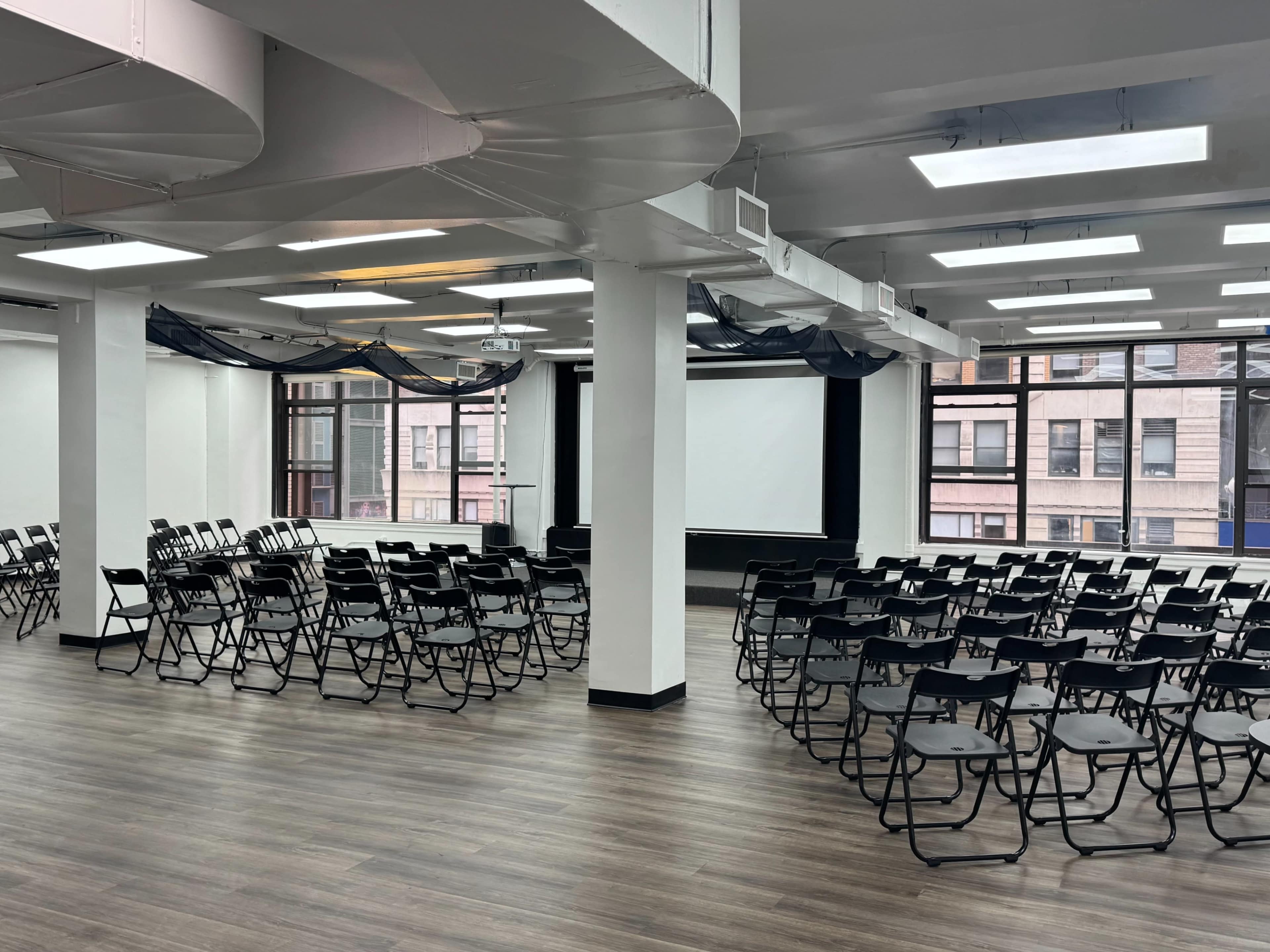 A spacious, well-lit room is set up with rows of black folding chairs facing a projection screen, with large windows providing a view of surrounding buildings.