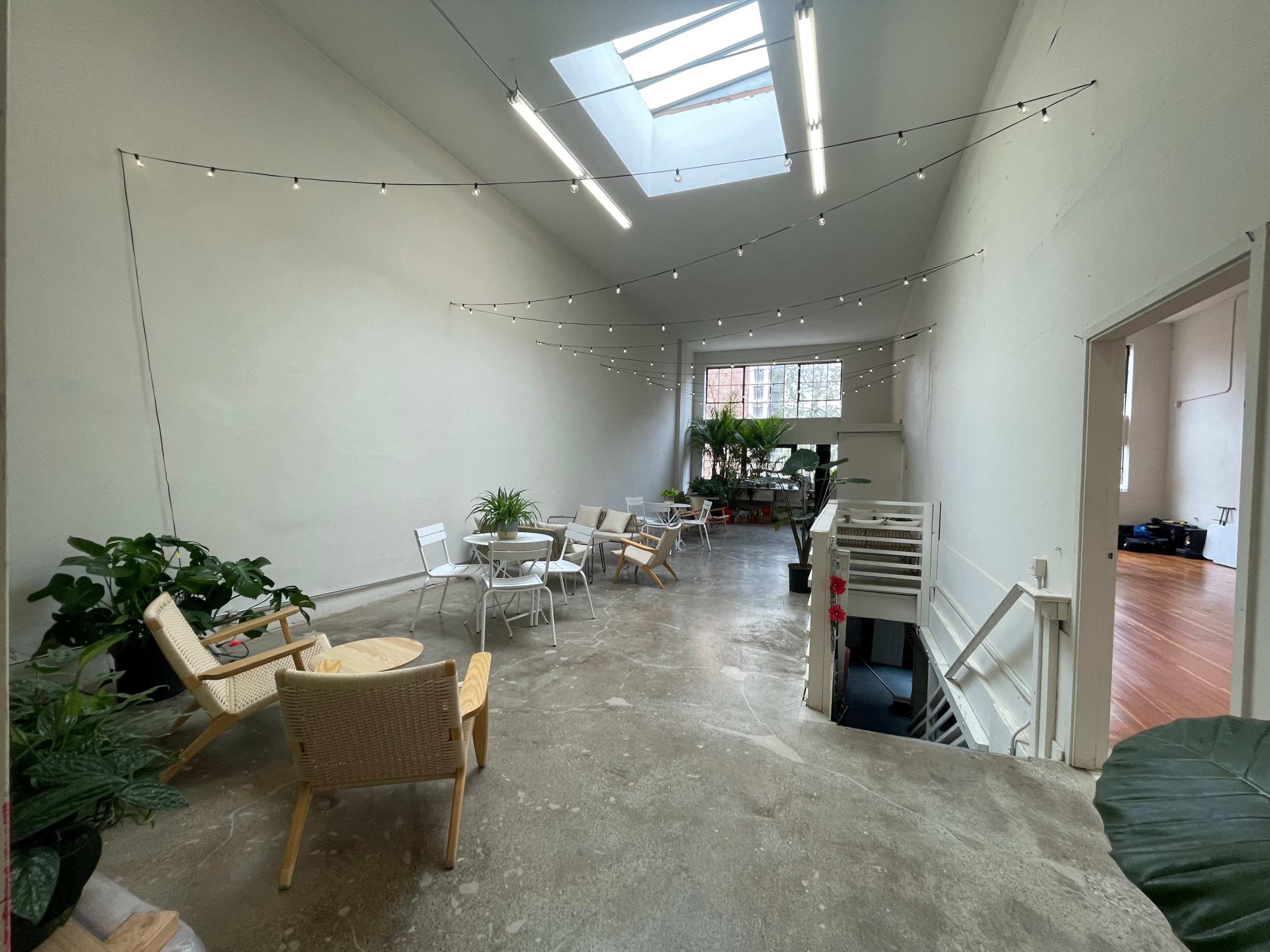 A spacious, well-lit interior with plants, seating arrangements, and string lights hanging from the ceiling.