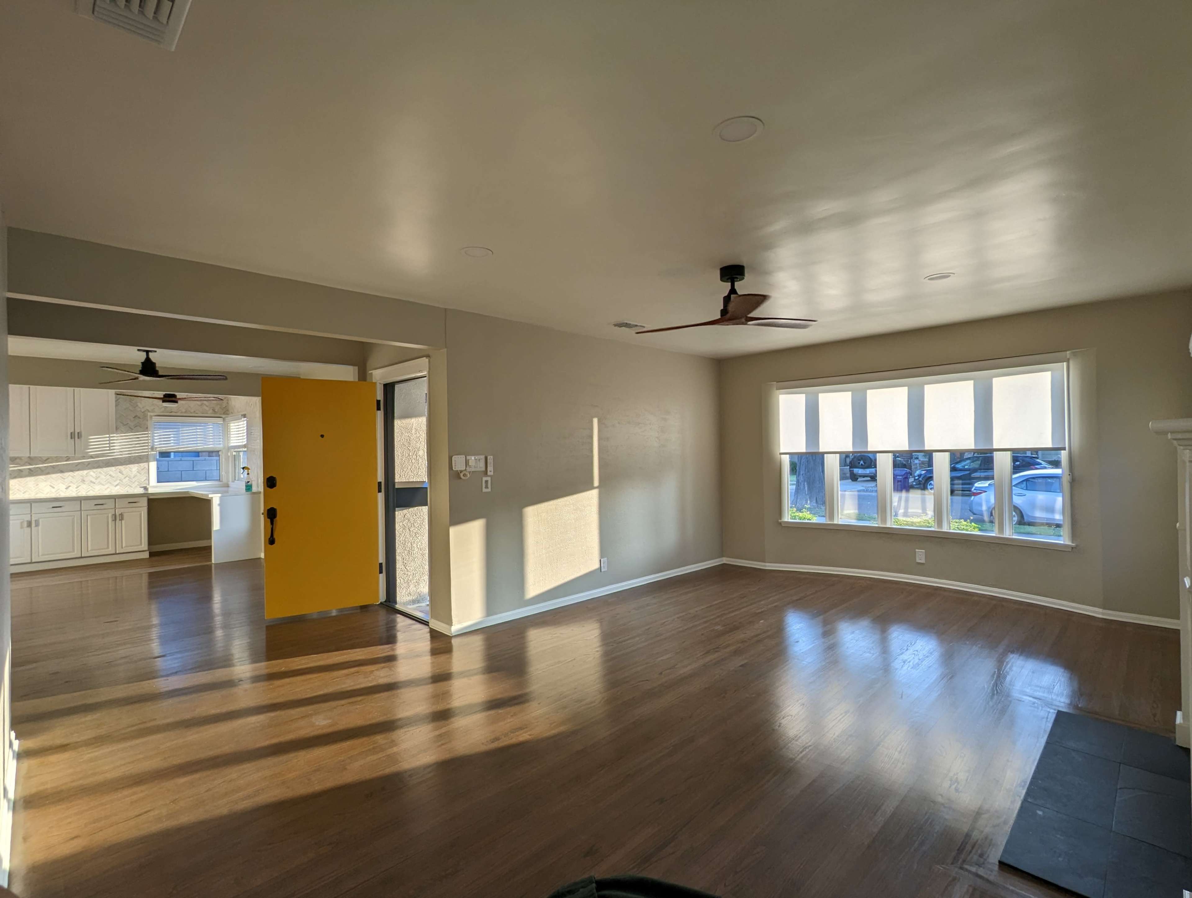 The image shows a spacious, light-filled living room with wooden floors and a view of a kitchen through an open doorway.