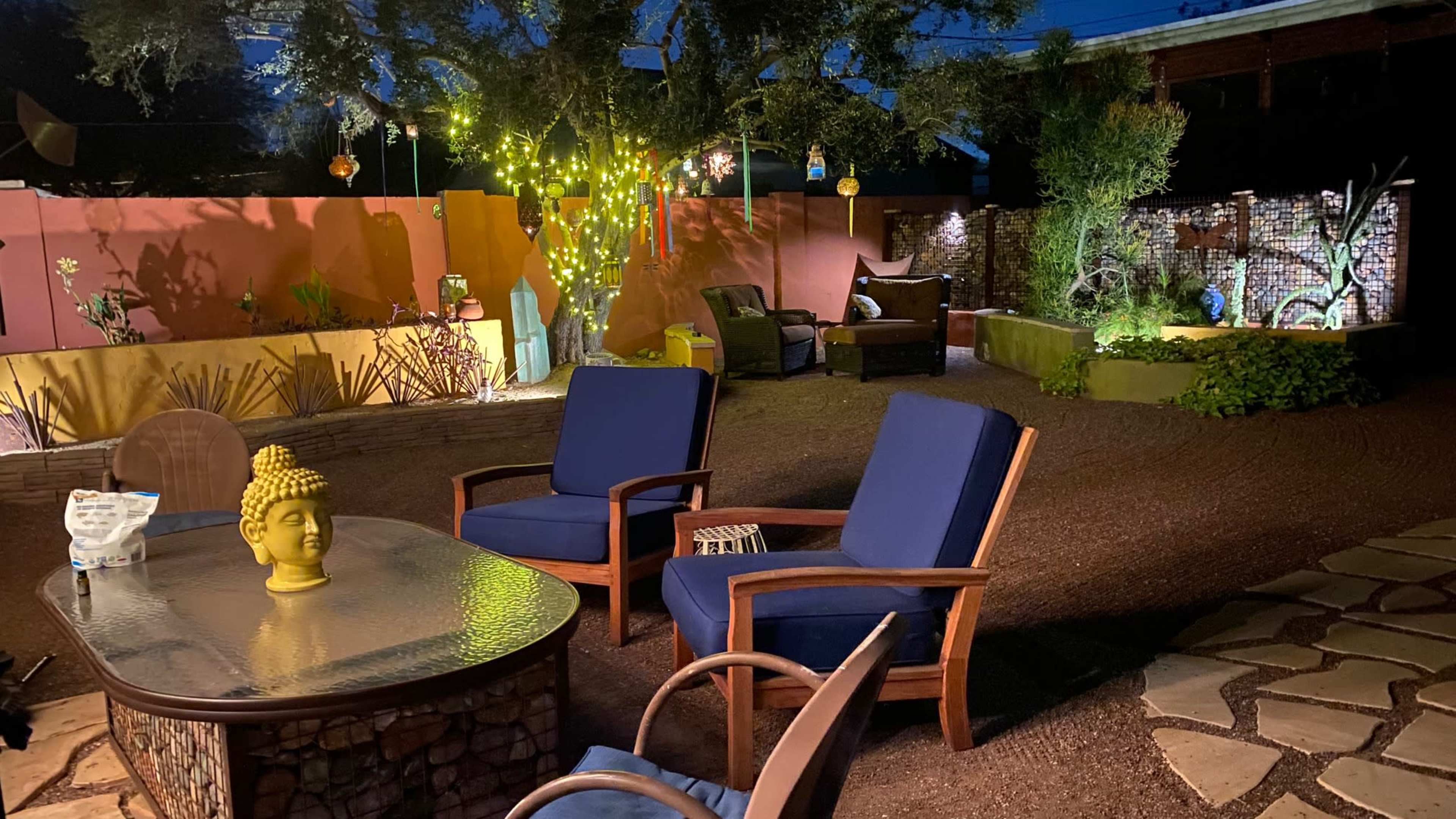 The image shows a landscaped outdoor patio at night, featuring a table with a Buddha statue and two blue chairs, surrounded by decorative lights and plants.
