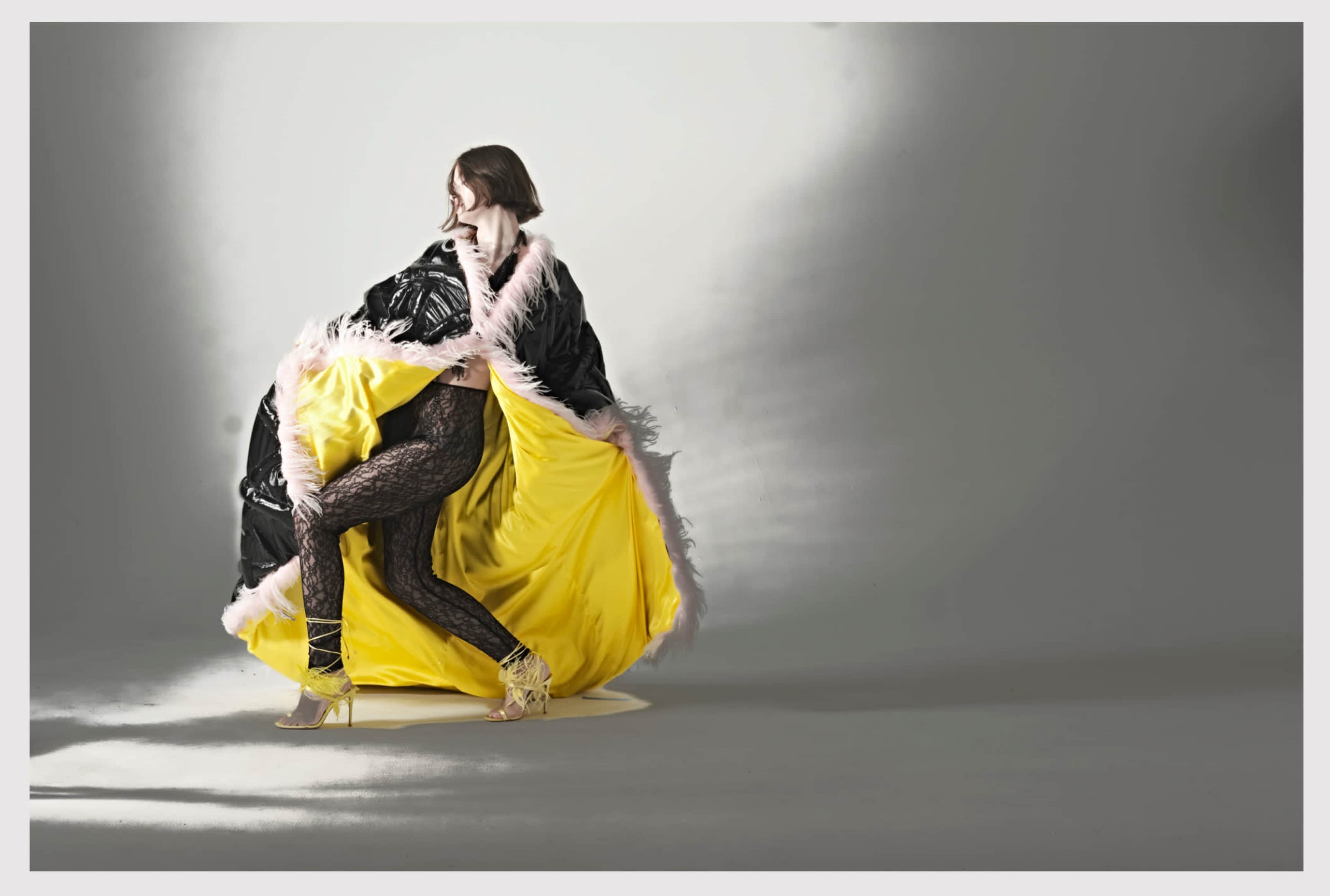 A person in a black and yellow patterned outfit stands against a gray backdrop, with a vibrant yellow lining and fluffy trim, while striking a dynamic pose.