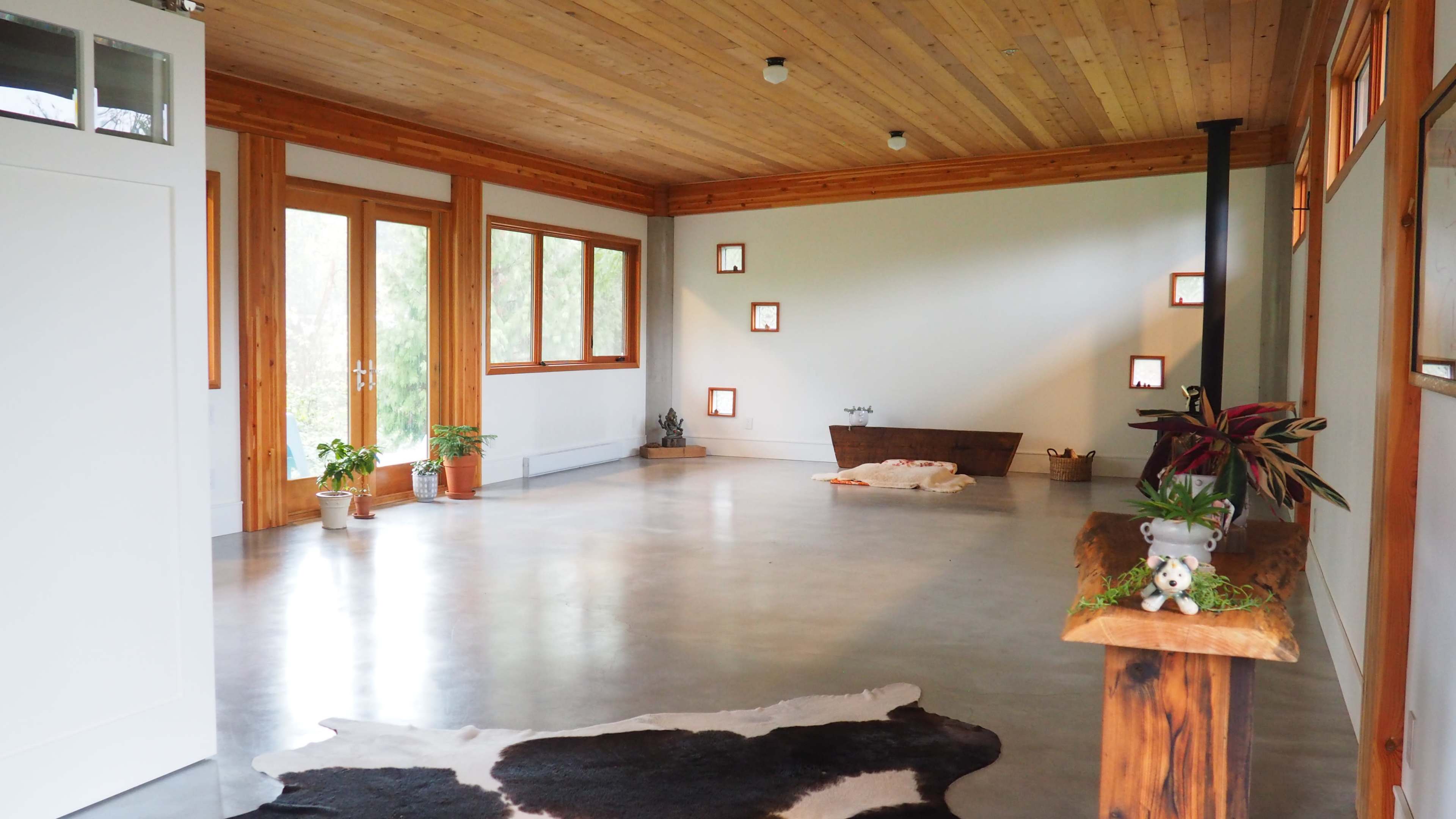 A spacious room with wooden ceilings and large windows, featuring a polished concrete floor, potted plants, and minimalist decor.