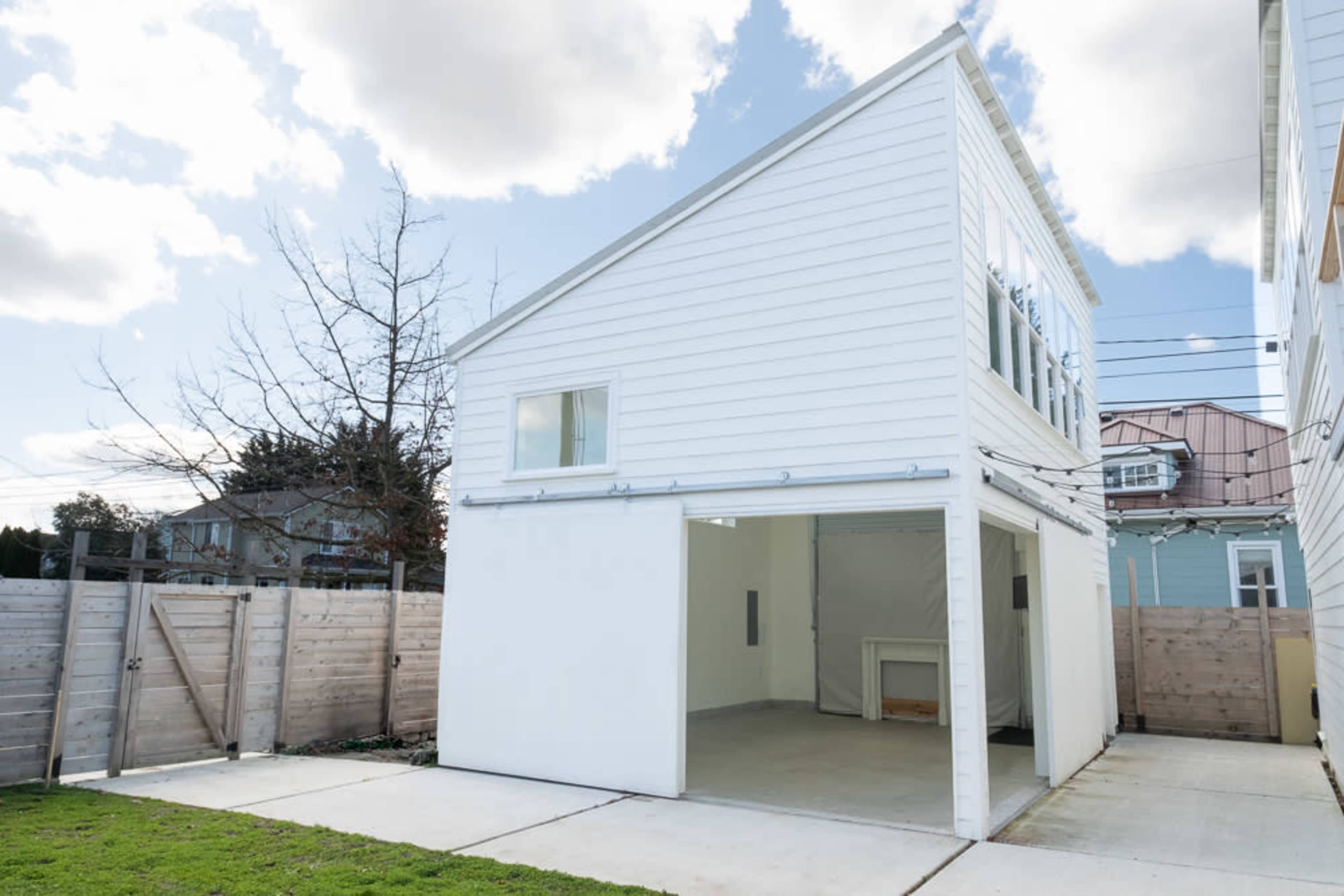 The image shows a modern, two-story white structure with large windows and an open garage area, set in an outdoor space with a grassy yard and a wooden fence.