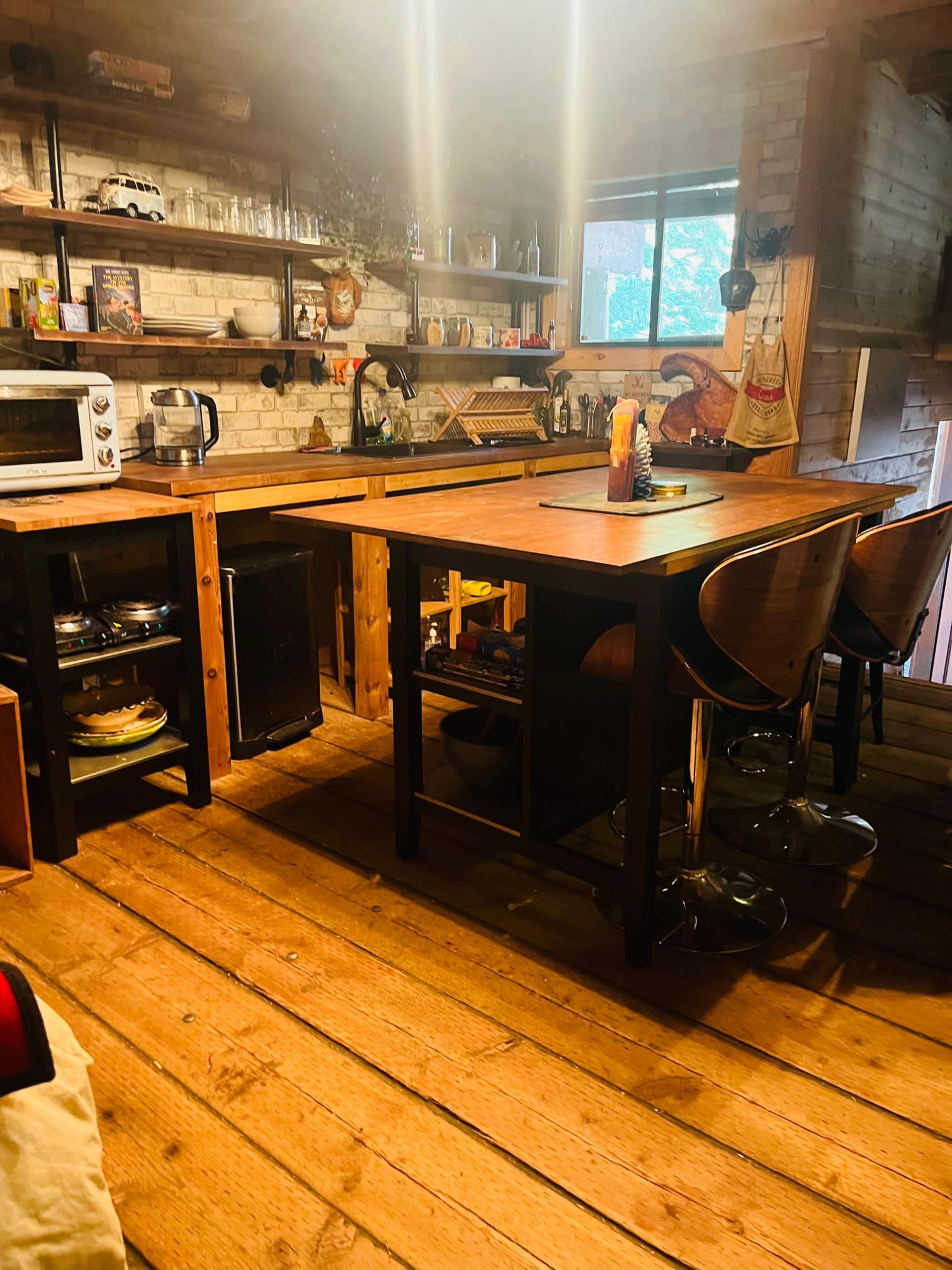 A wooden kitchen with a large table, bar stools, and shelves filled with kitchenware and appliances.