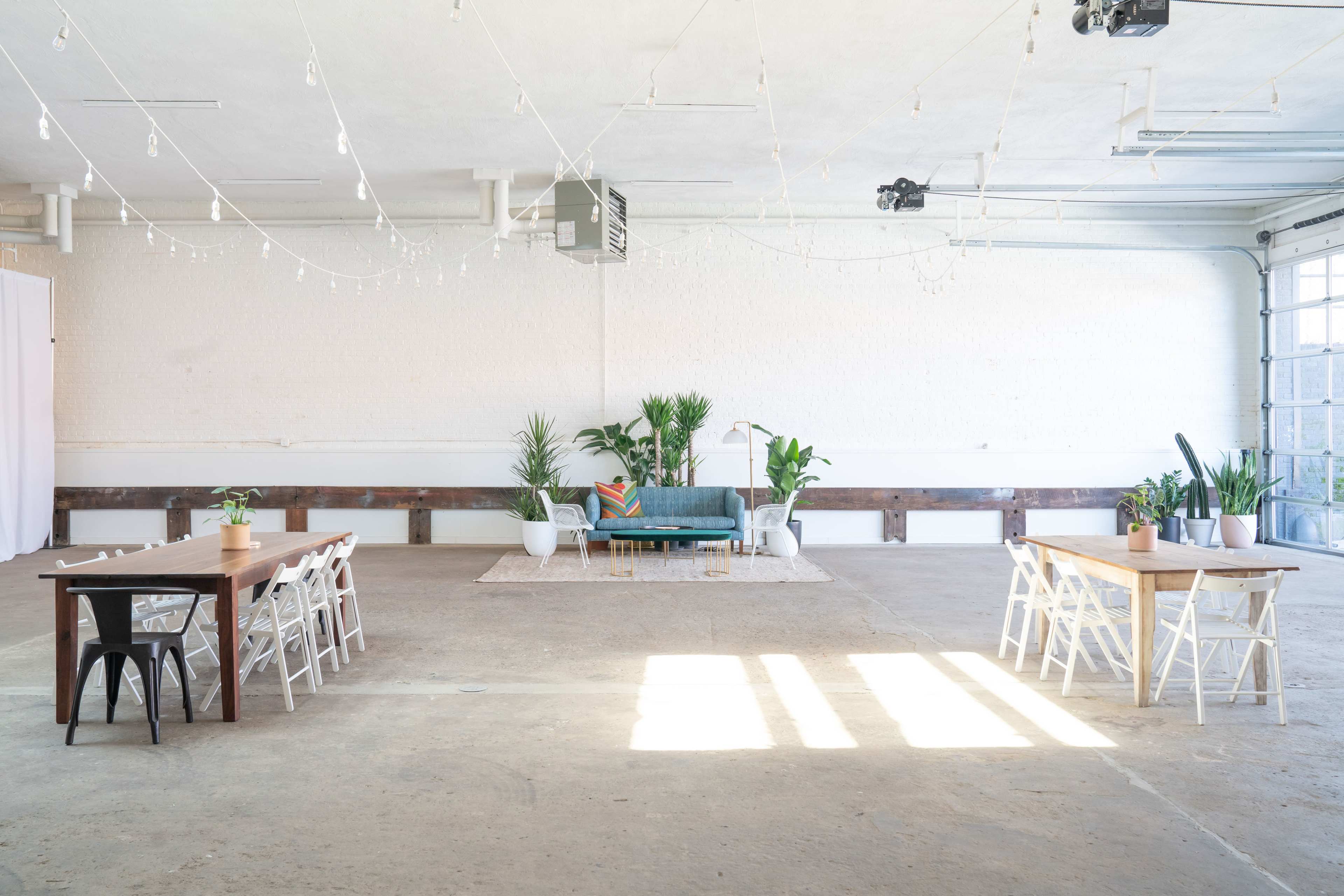 A spacious, well-lit room features a teal couch and several plants, with two tables set up on either side.
