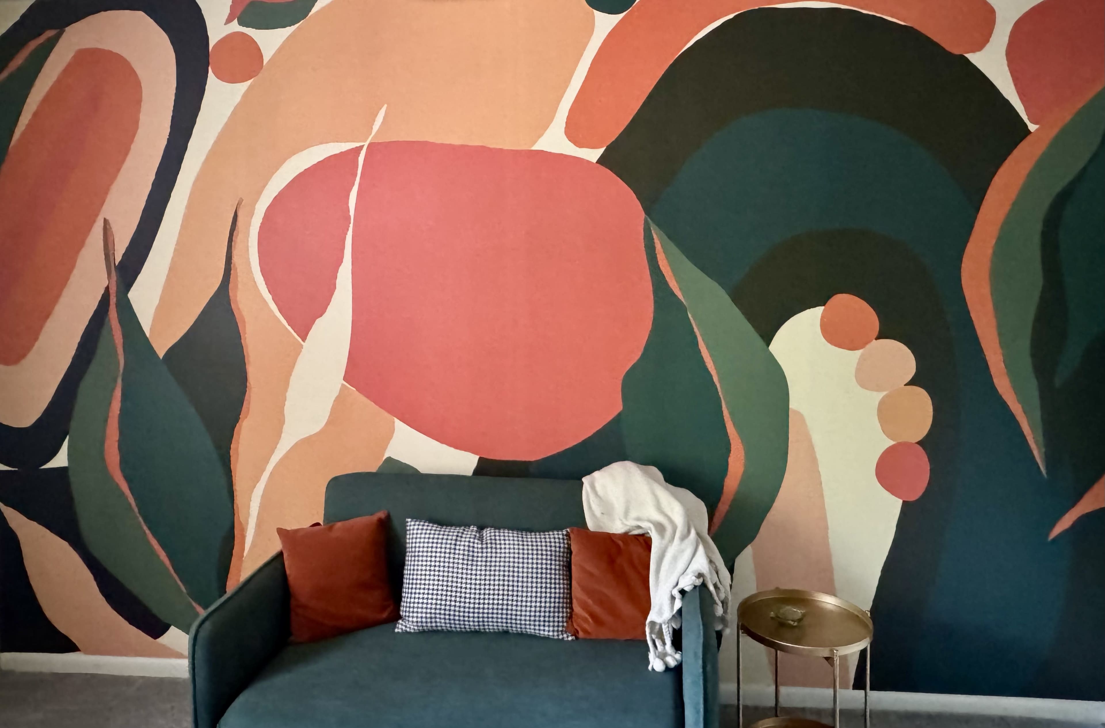 The image shows a teal sofa with orange and patterned pillows in front of a colorful abstract mural featuring organic shapes and bold colors on the wall.