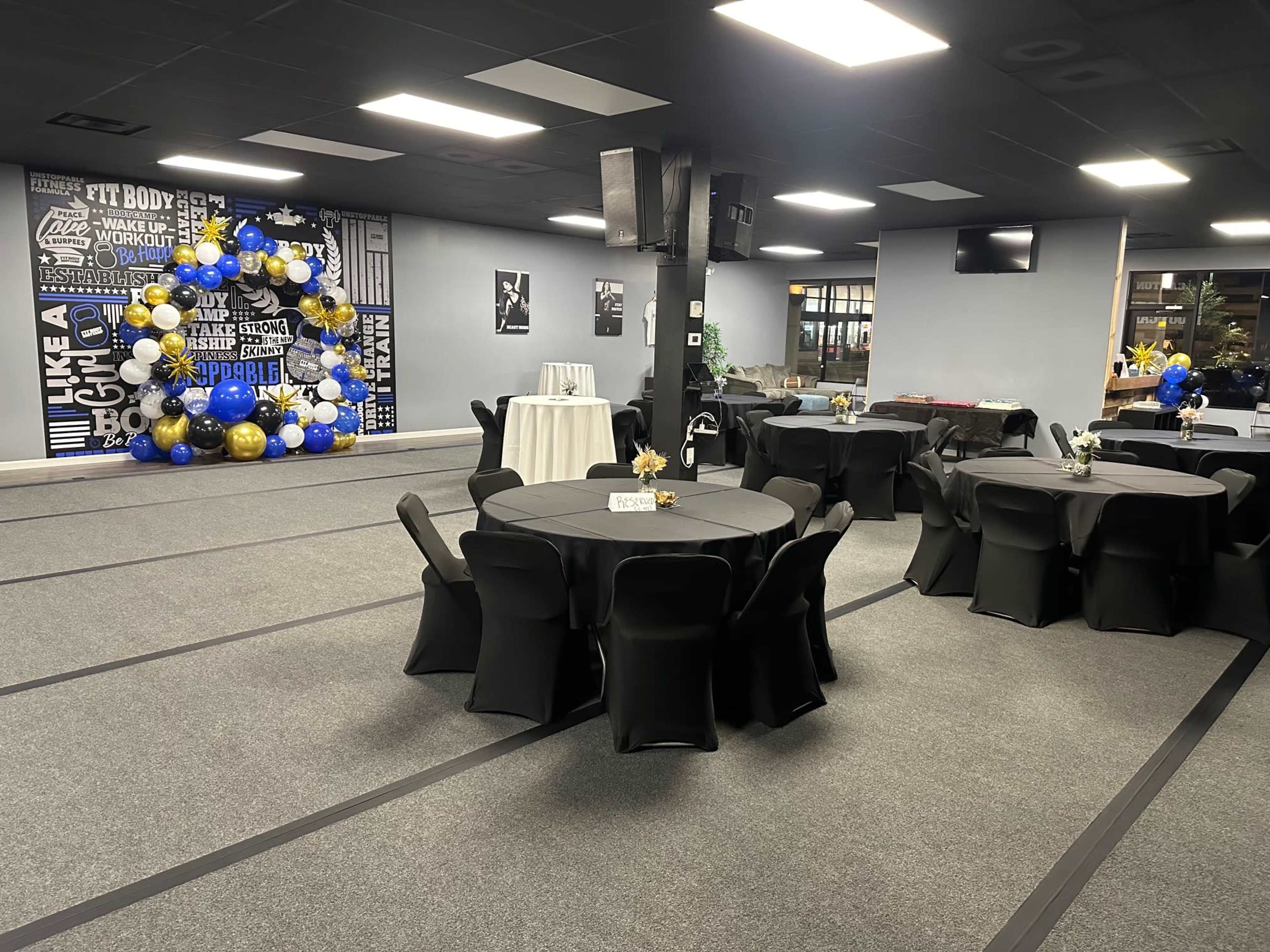 The image shows a decorated event space with round tables covered in black cloth, a floral balloon arch backdrop, and various centerpieces.
