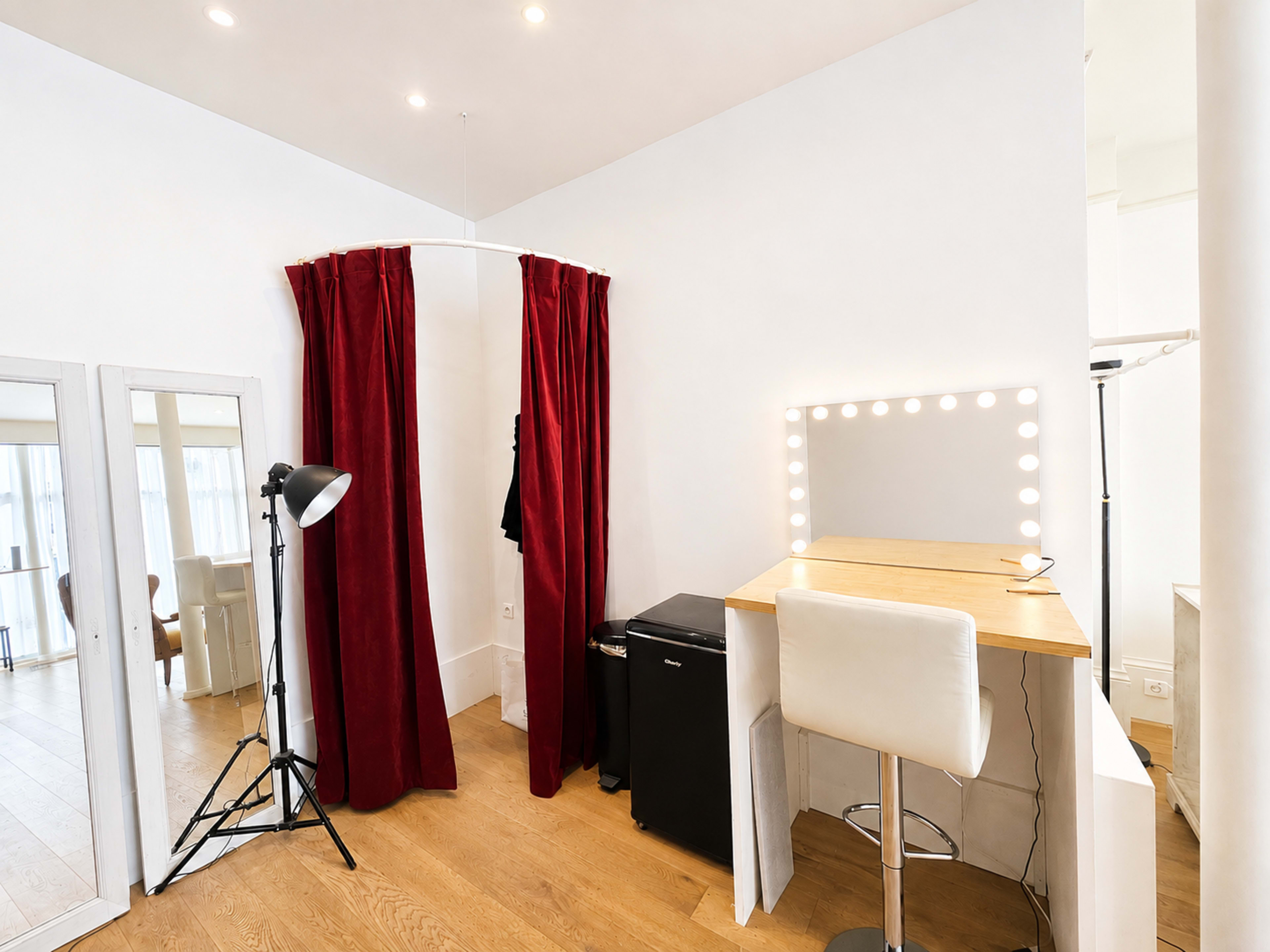 The image shows a small dressing area featuring a wooden vanity with a mirror surrounded by lights, a chair, a mini-fridge, a photography light stand, and a pair of red curtains.