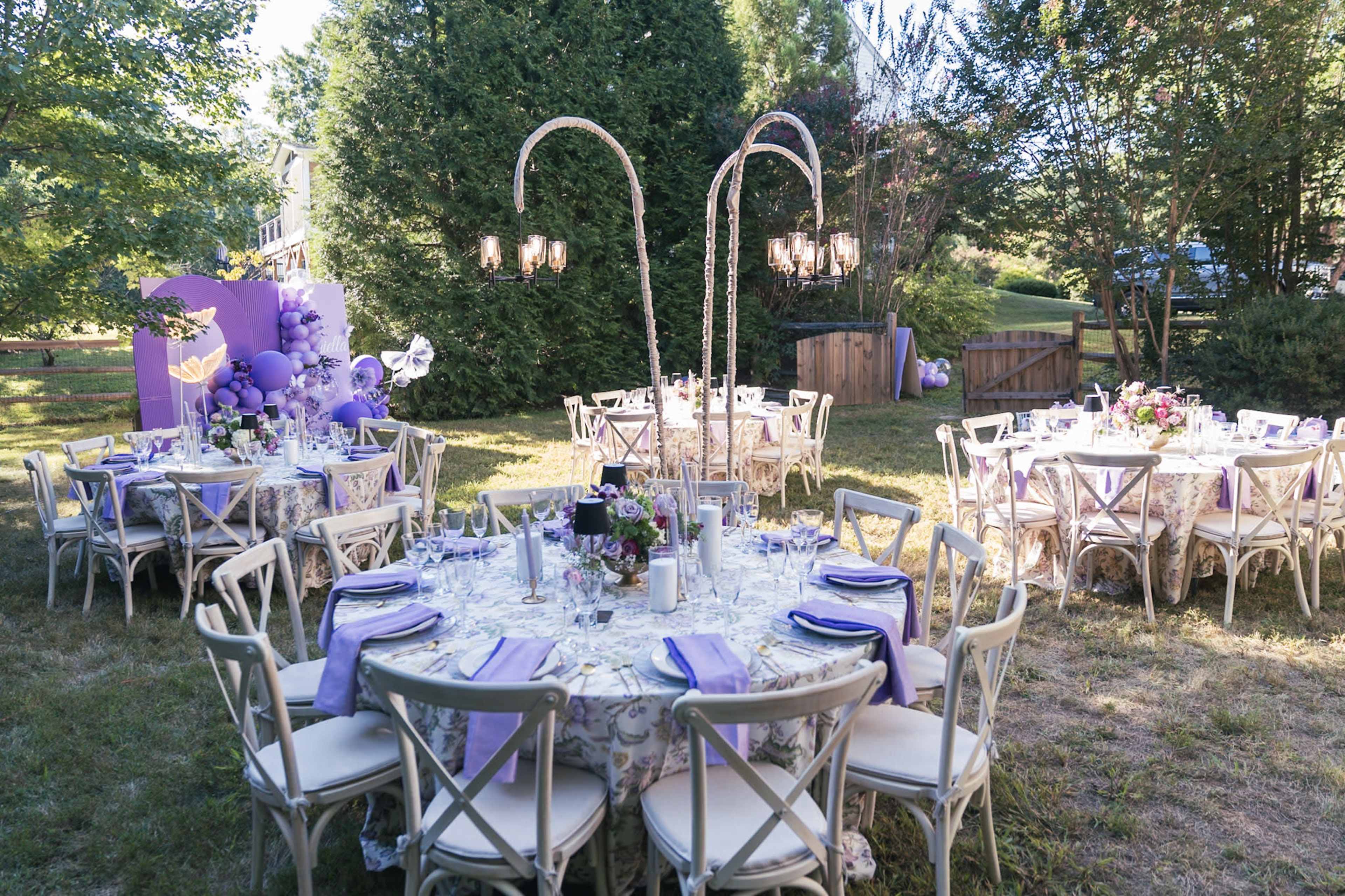 The image shows an outdoor dining area set for an event, featuring multiple round tables with elegant table settings under trees.