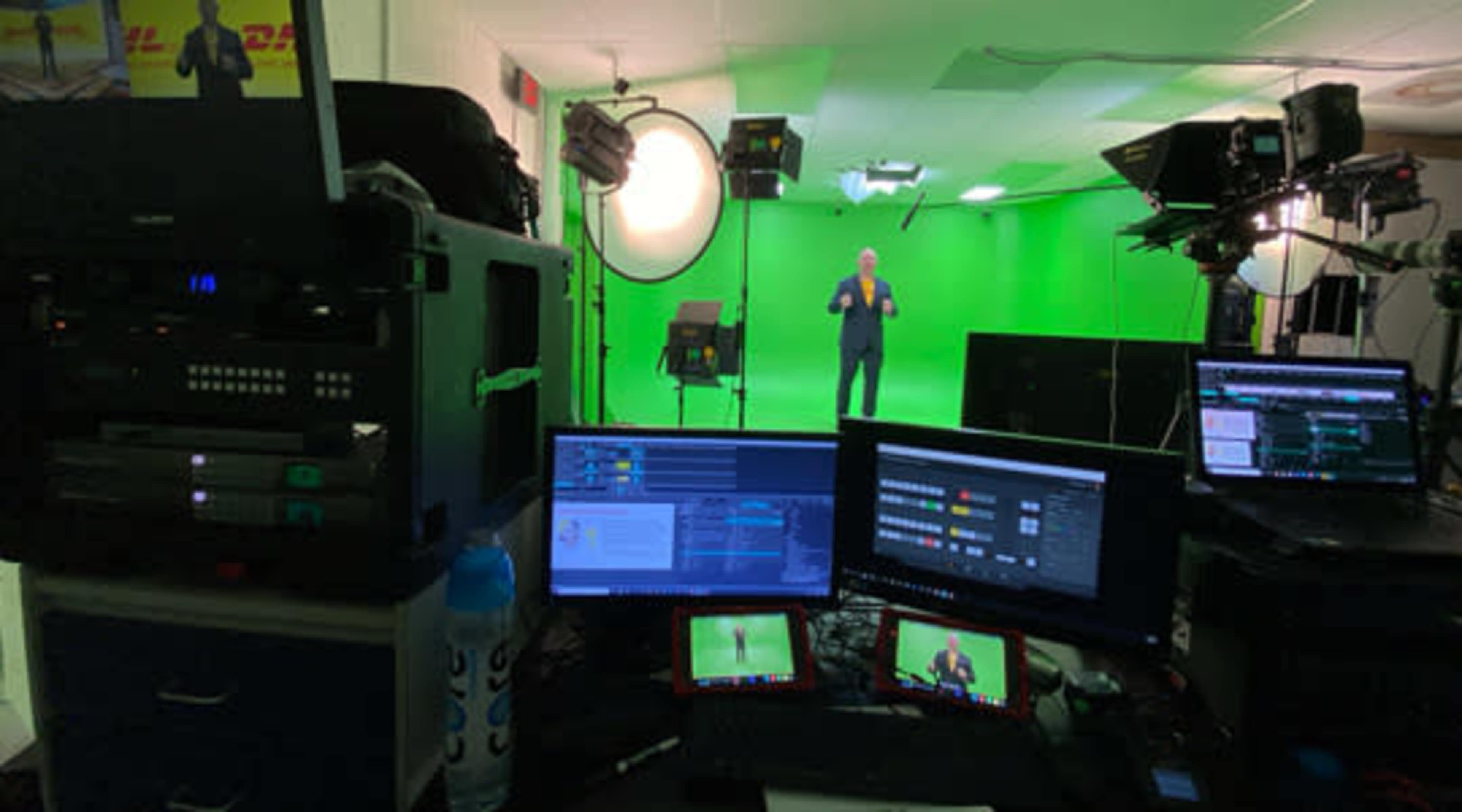 The image shows a green screen studio setup with multiple computers, monitors, and lighting equipment, while a person stands in front of the green backdrop.