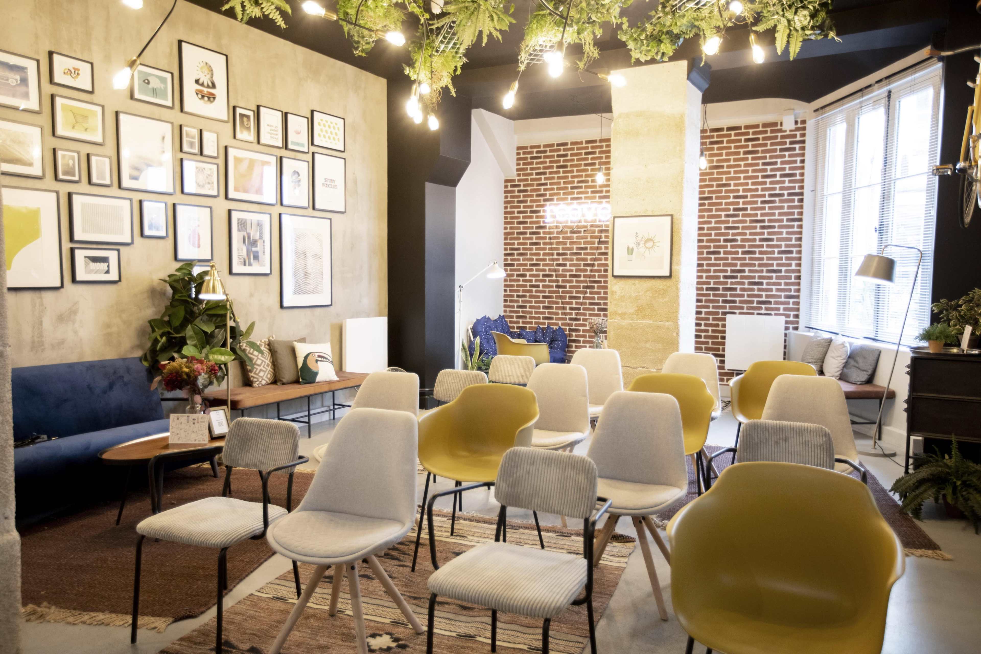 The image shows a modern event space with an arrangement of chairs facing a wall decorated with framed art and greenery, set against a backdrop of exposed brick.