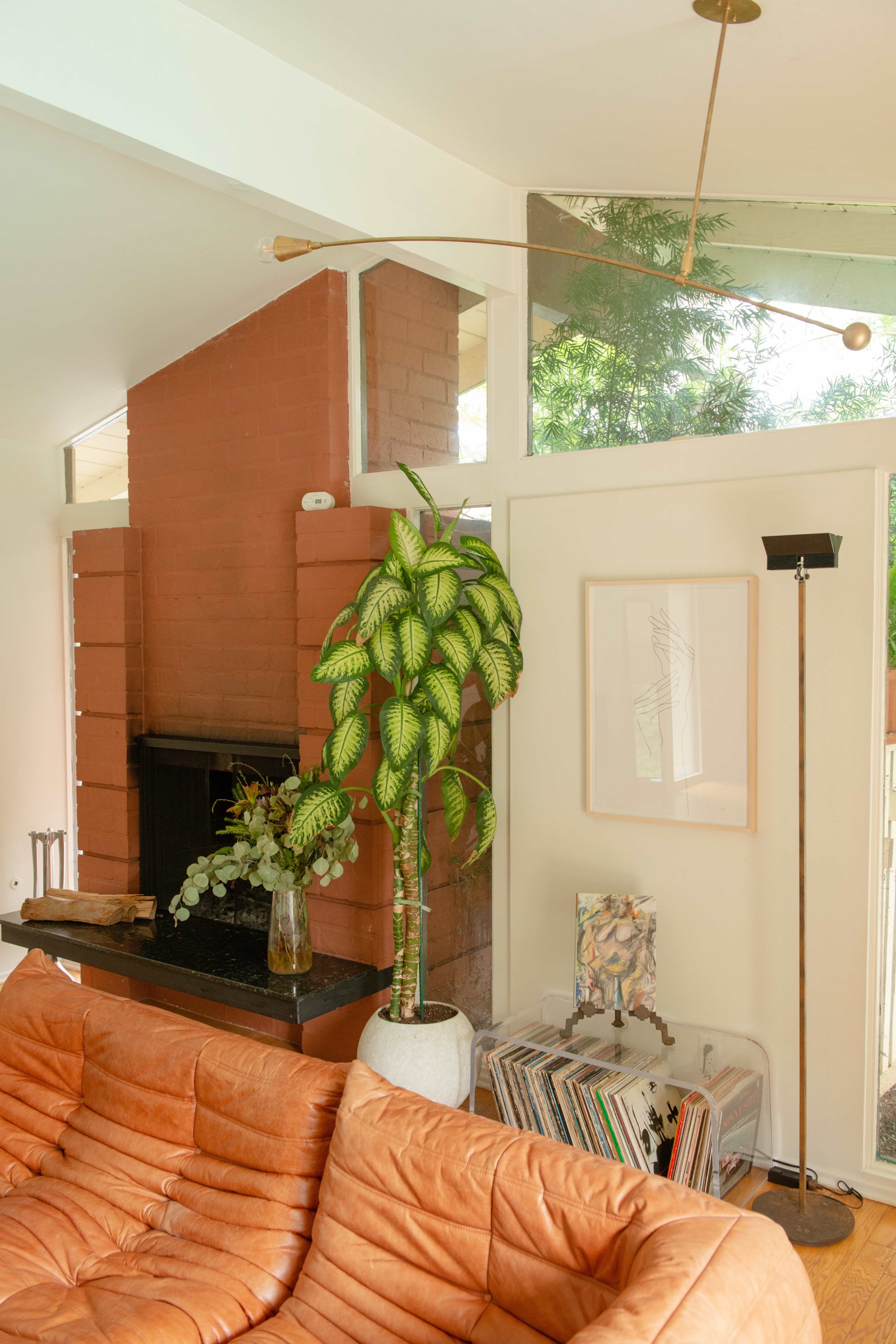 A well-lit living room featuring a brown leather couch, a tall potted plant, a brick fireplace, and a collection of magazines on a clear shelf.