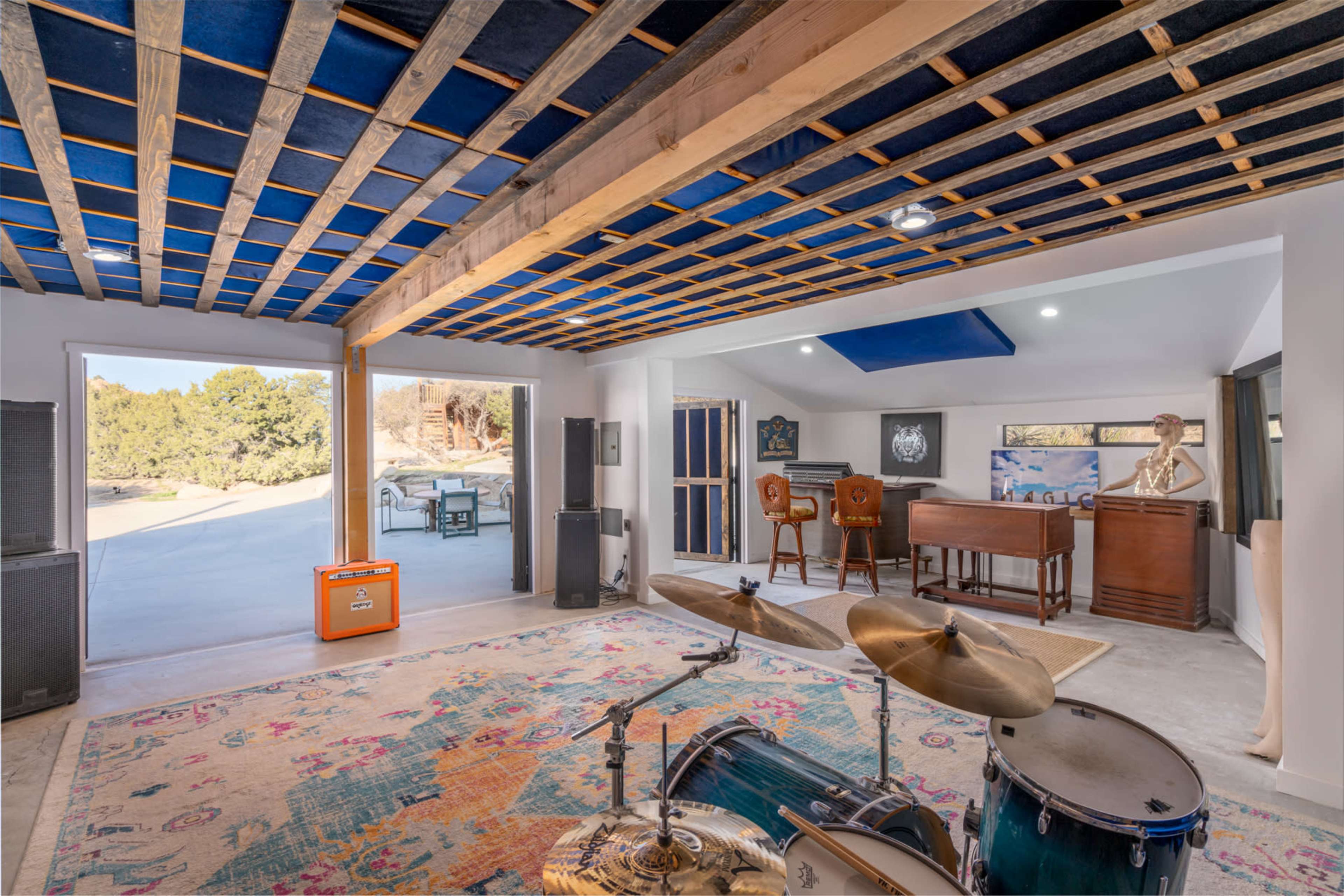 A spacious room with a drum set in the foreground and a cozy seating area in the background, featuring a piano and artwork.