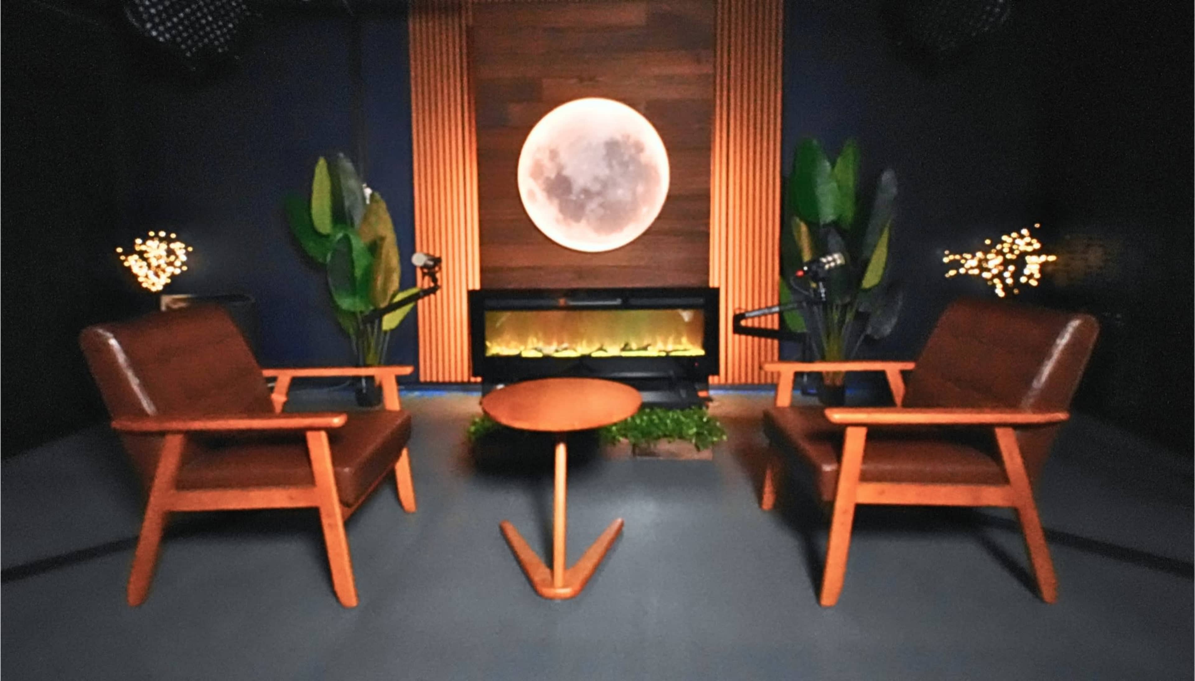 The image features a cozy indoor setting with two wooden chairs facing a circular wall art resembling the moon, flanked by artificial plants and a fireplace in a dimly lit room.