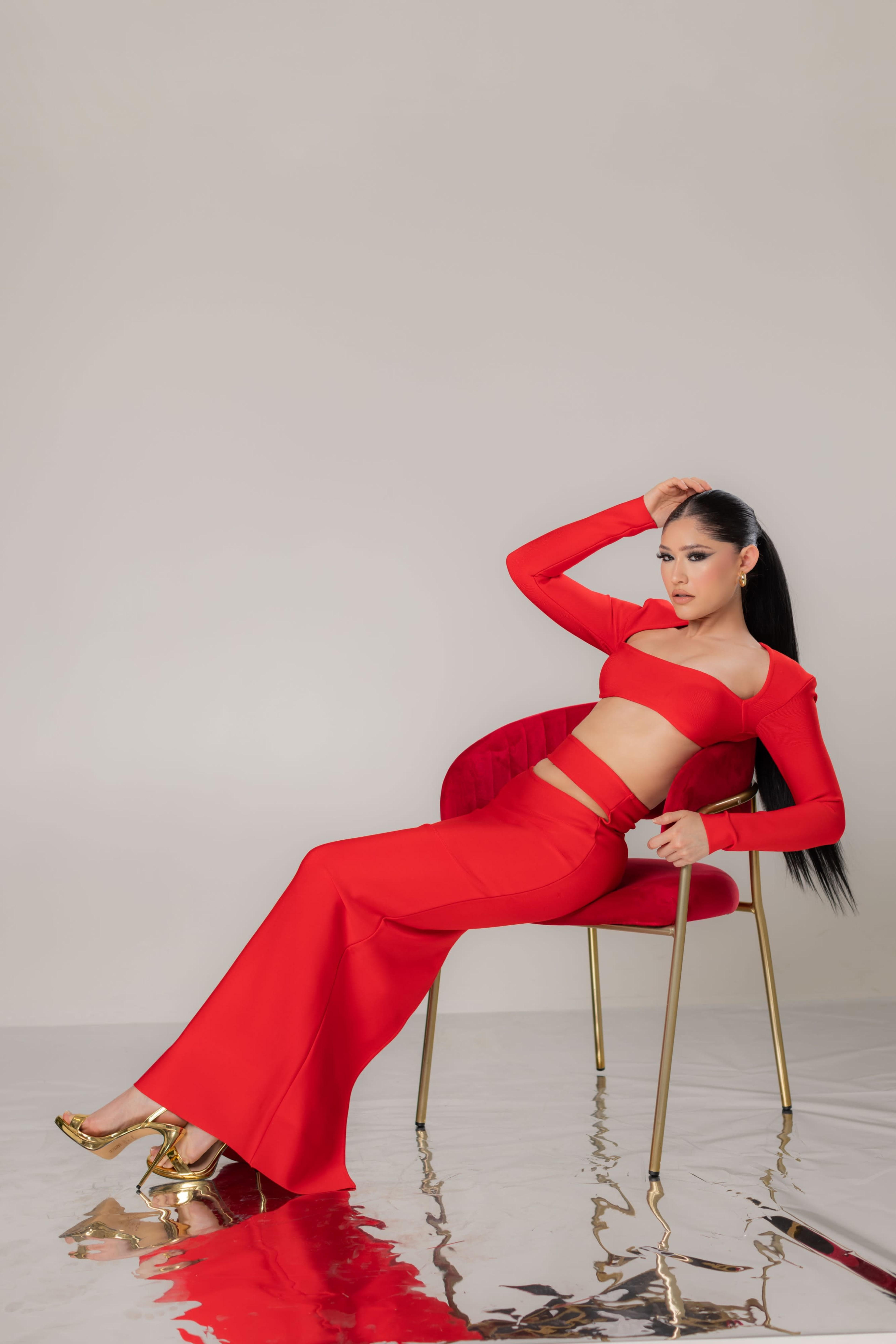 A person in a red two-piece outfit sits gracefully on a gold-accented chair against a neutral background.