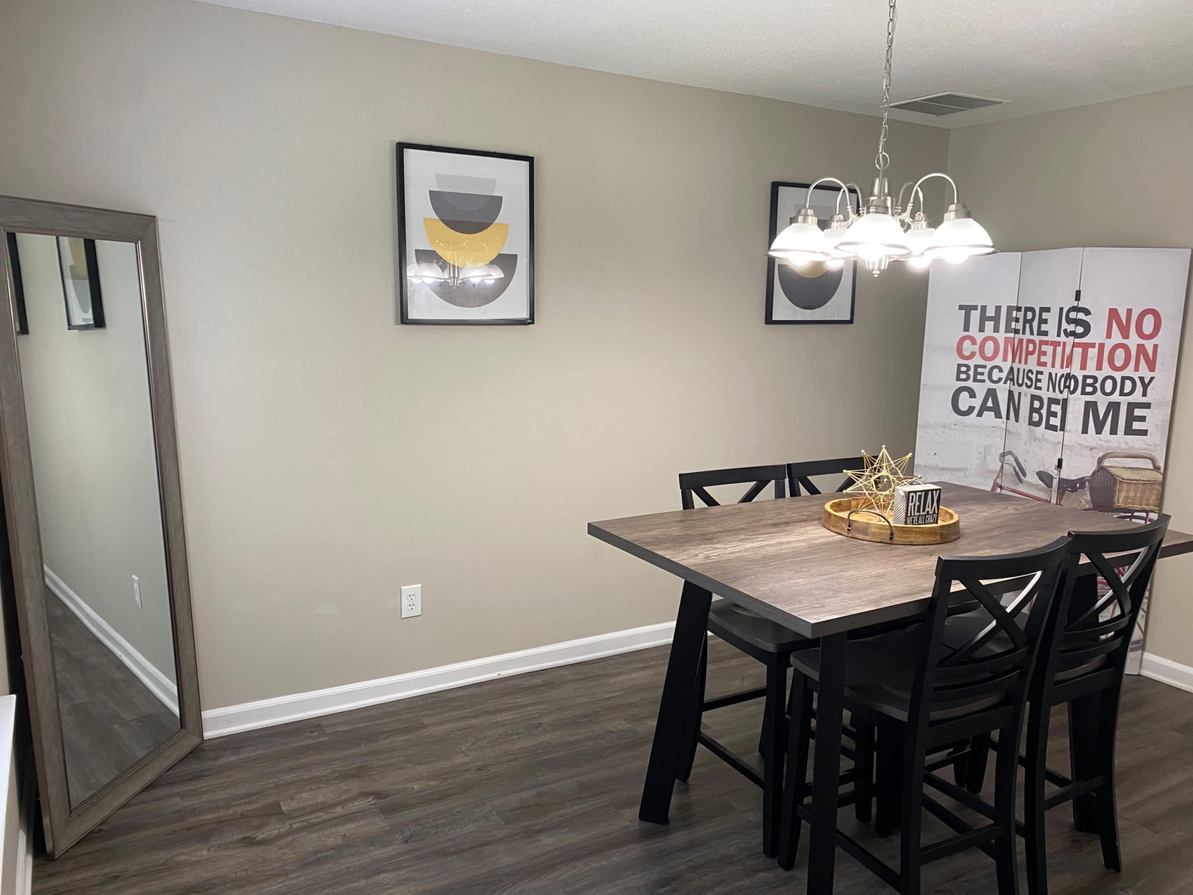 A dining area features a wooden table with black chairs, a large decorative mirror, and wall art including a motivational sign and framed prints.