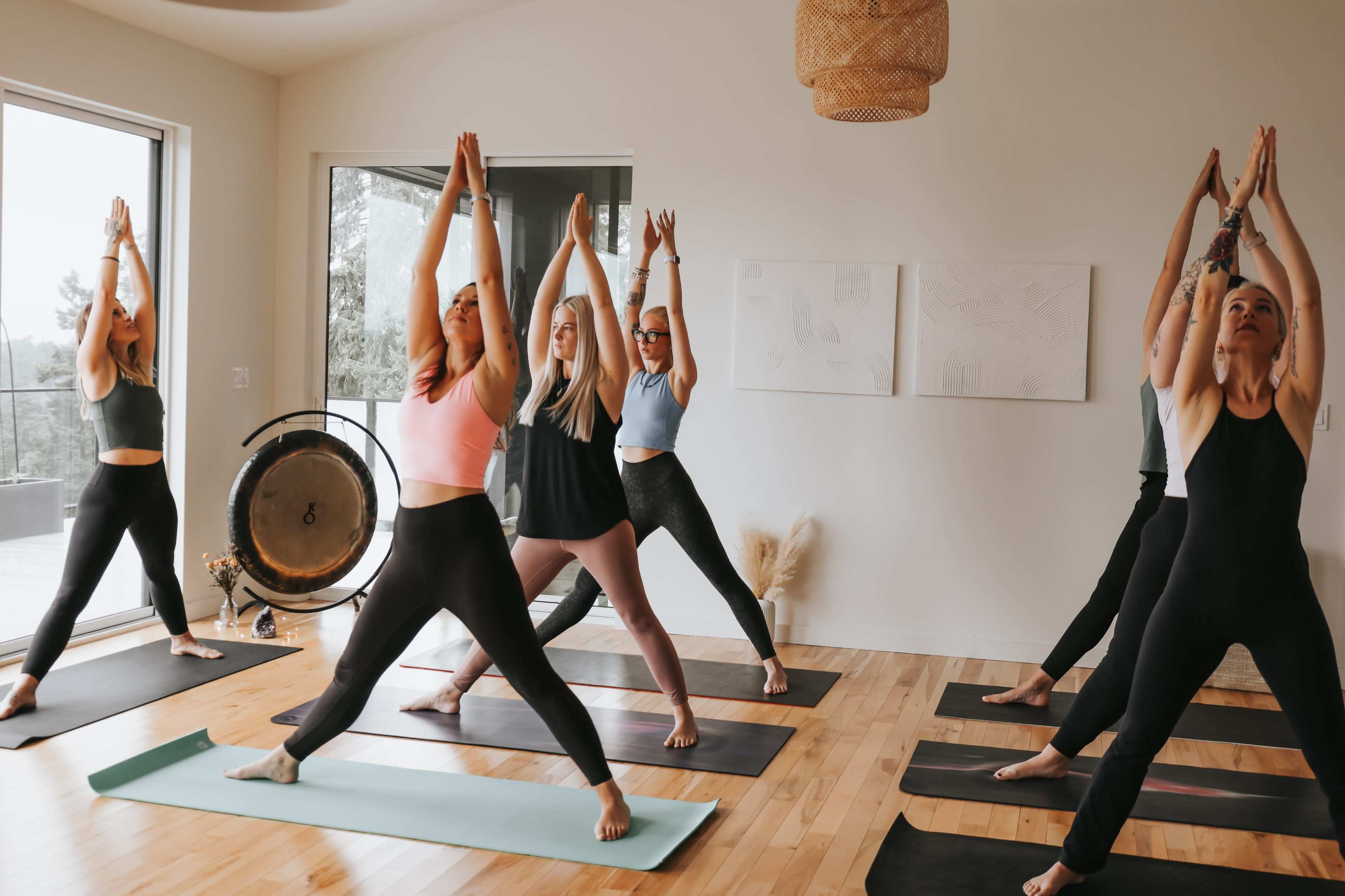 A group of six women in yoga attire practices a standing pose in a well-lit studio with wooden floors and large windows.