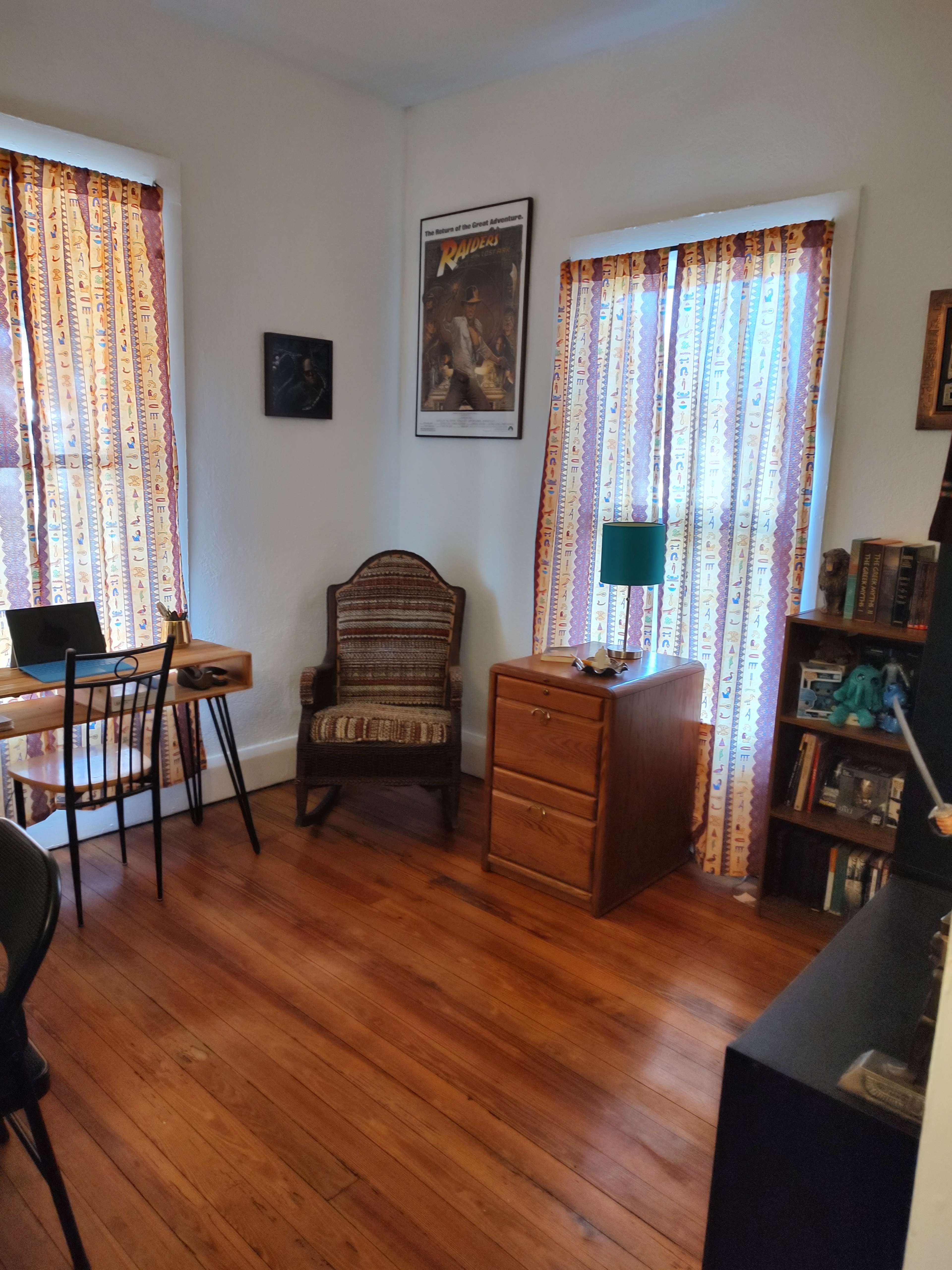 A cozy room with two windows draped in patterned curtains, a wooden chair, a small desk, a lamp on a nightstand, and a bookshelf against the wall.