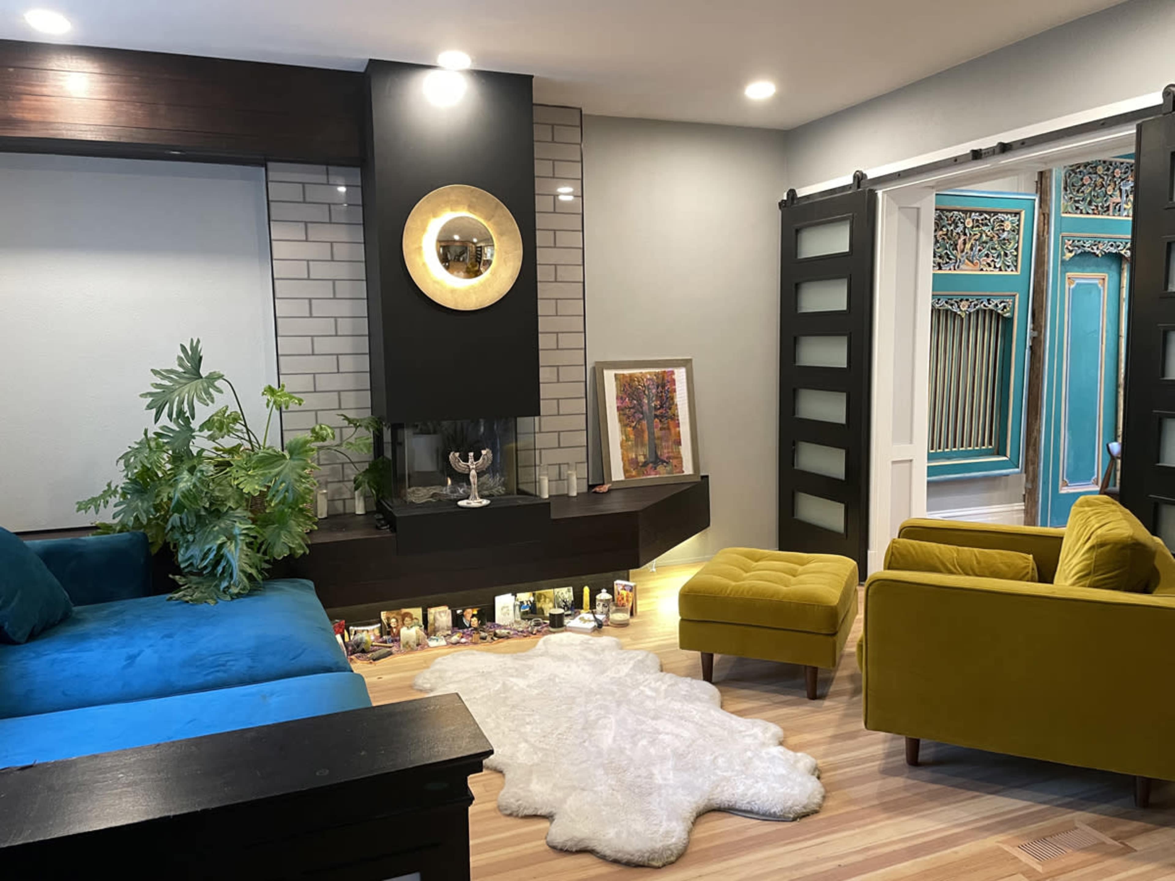 The image shows a modern living room with a blue sofa, two yellow armchairs, a white fur rug, and a decorative fireplace surrounded by framed artwork and plants.