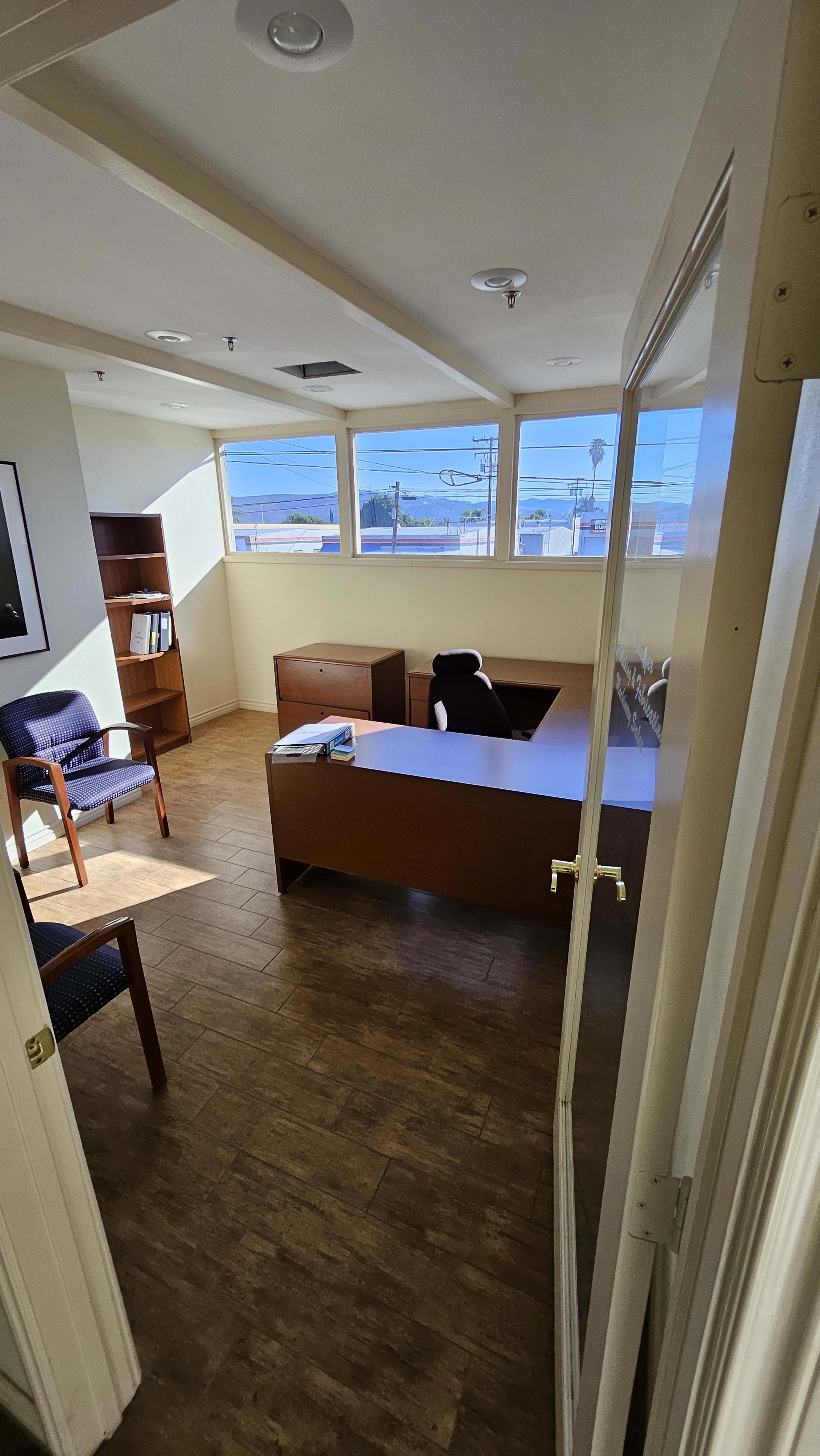 An office space with a desk, a chair, and a bookshelf, featuring windows that provide a view of the outdoors.