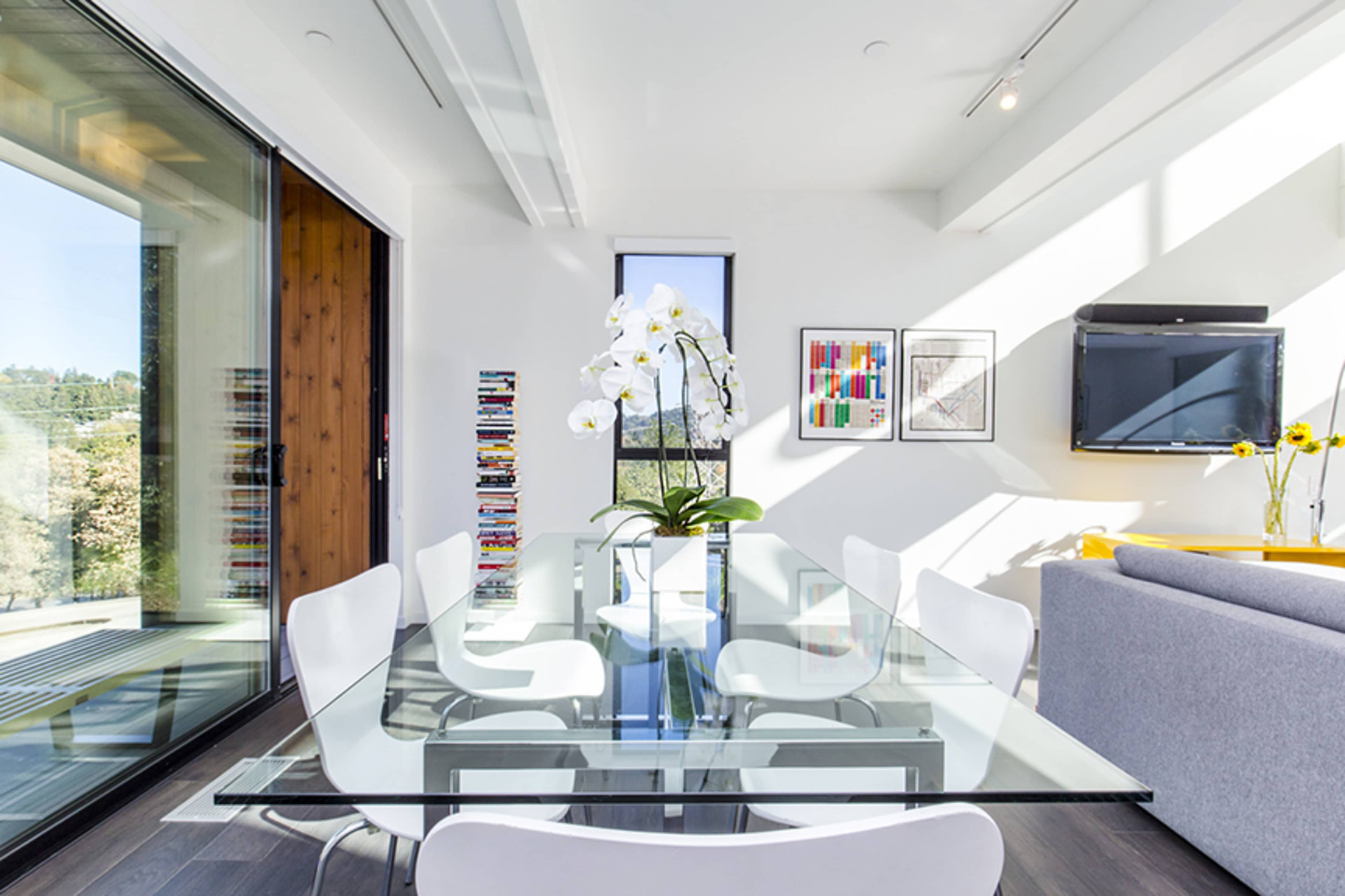 A modern dining area features a glass table surrounded by white chairs, with a large orchid centerpiece and a wall-mounted television.