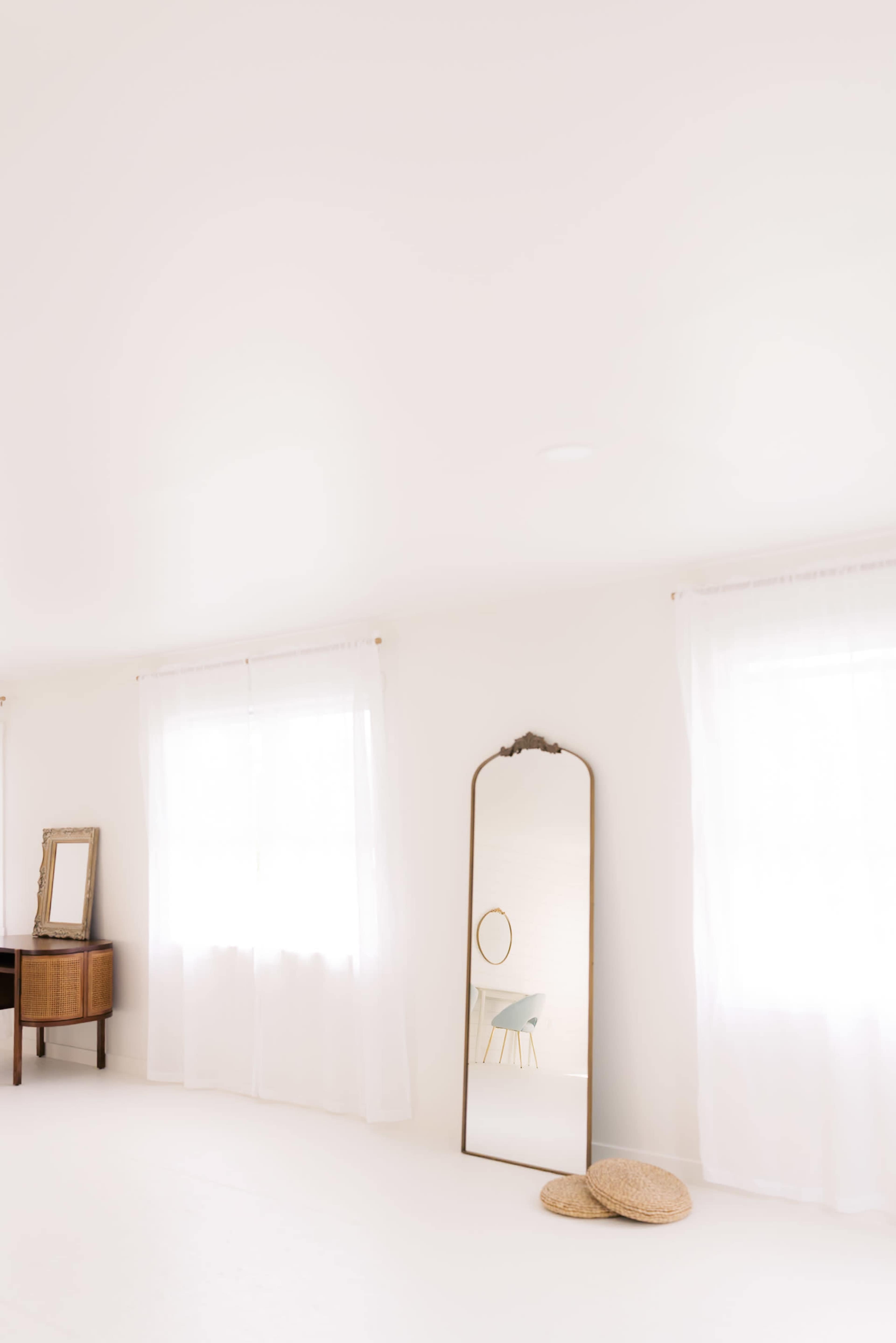 The room features large windows with sheer curtains, a standing mirror, a small round mirror on the wall, a wooden dresser, and two circular woven mats on the floor.