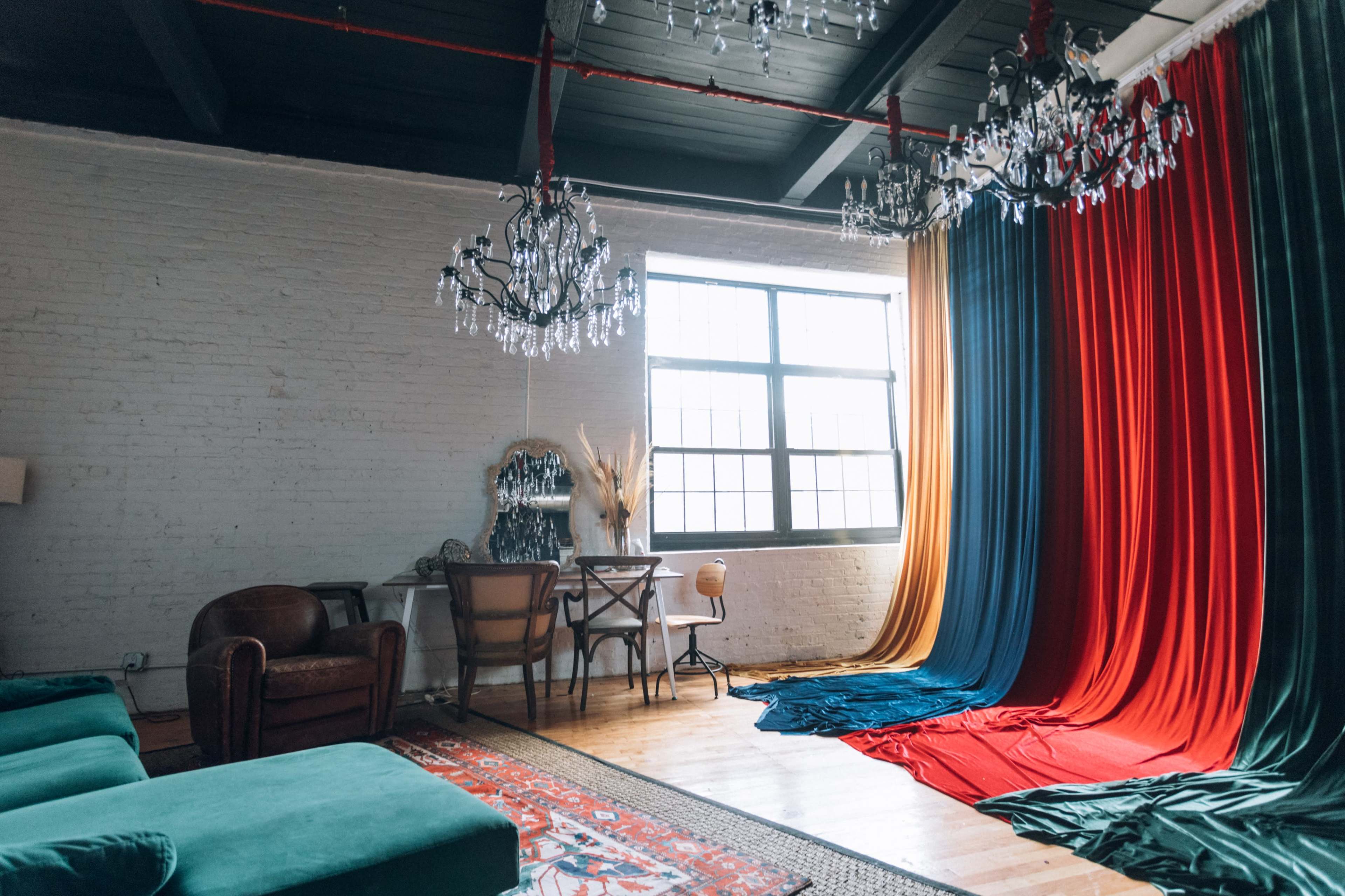 A brightly lit room features a large window, two chandeliers, and colorful fabric drapes in red, blue, and orange alongside a vintage armchair and a small dining table.