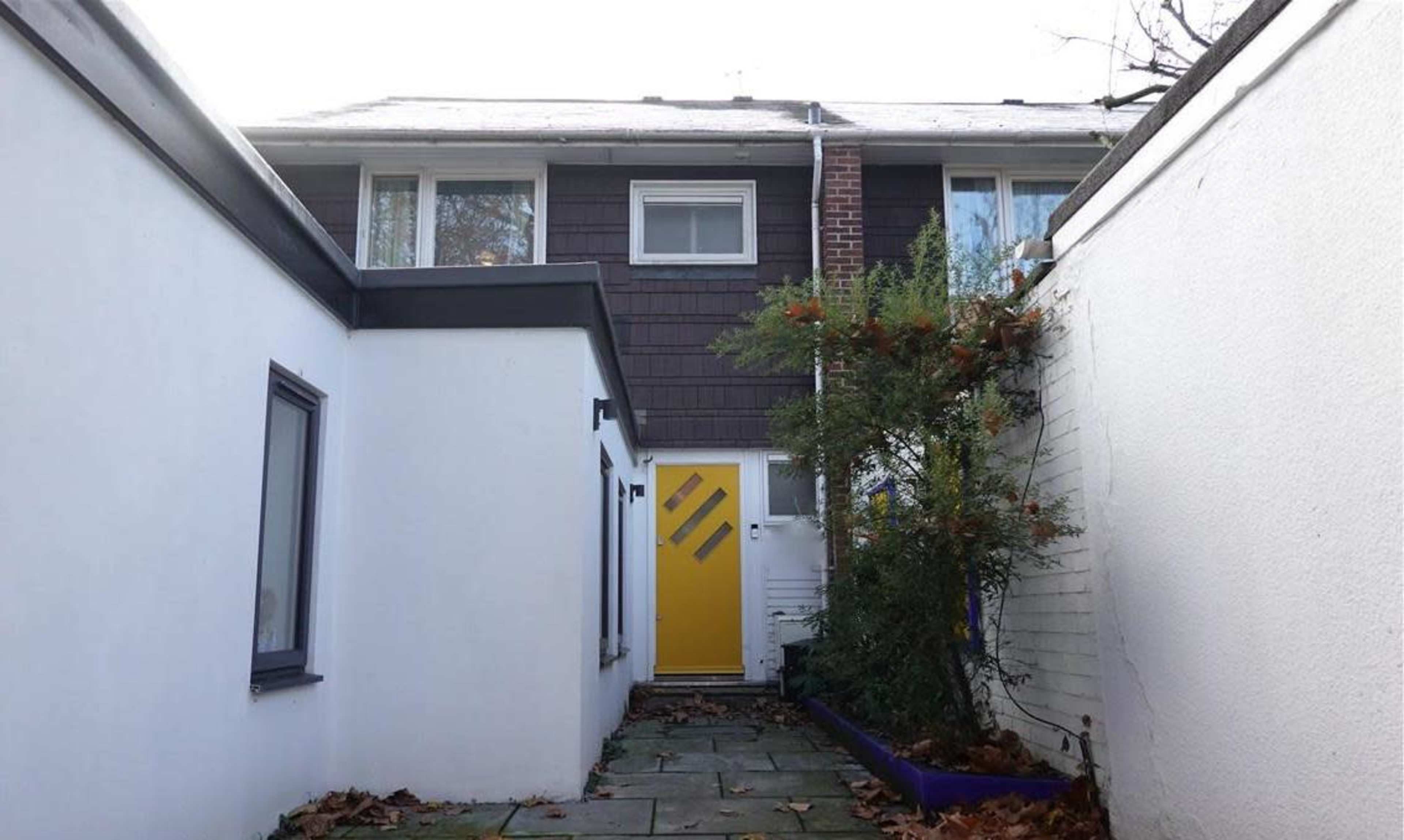 A narrow pathway leading to a yellow door of a house, flanked by white walls and a small garden area.