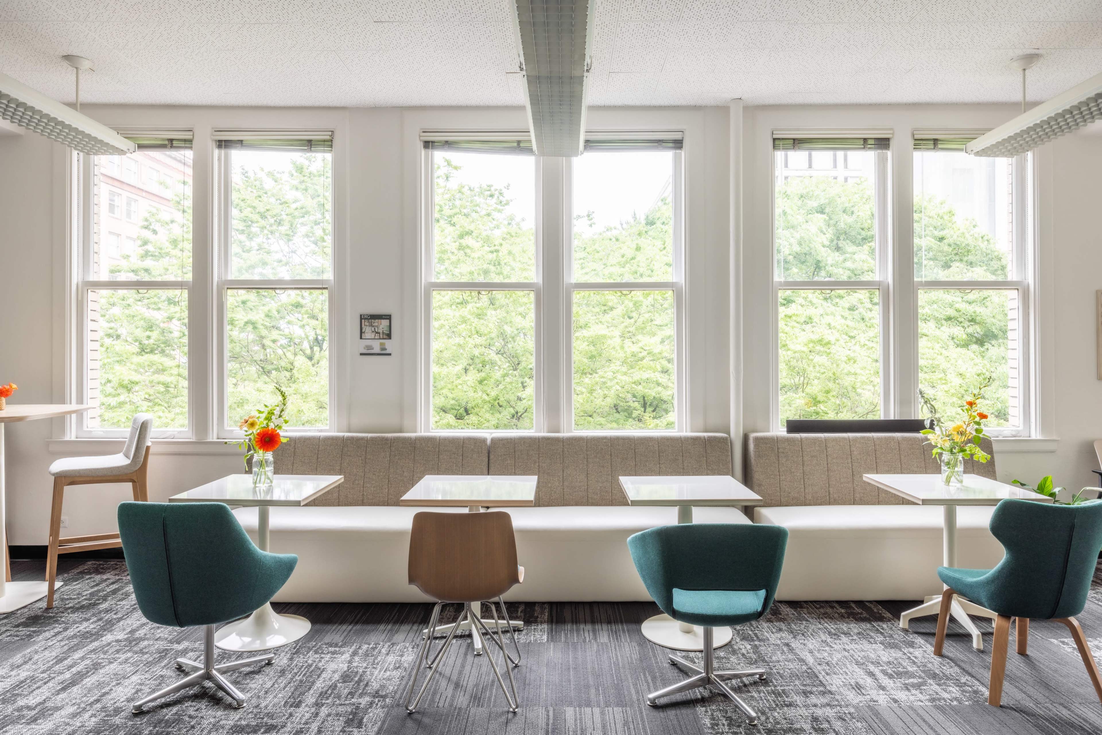 The image shows a modern café with several tables and colorful chairs arranged near large windows that overlook greenery.