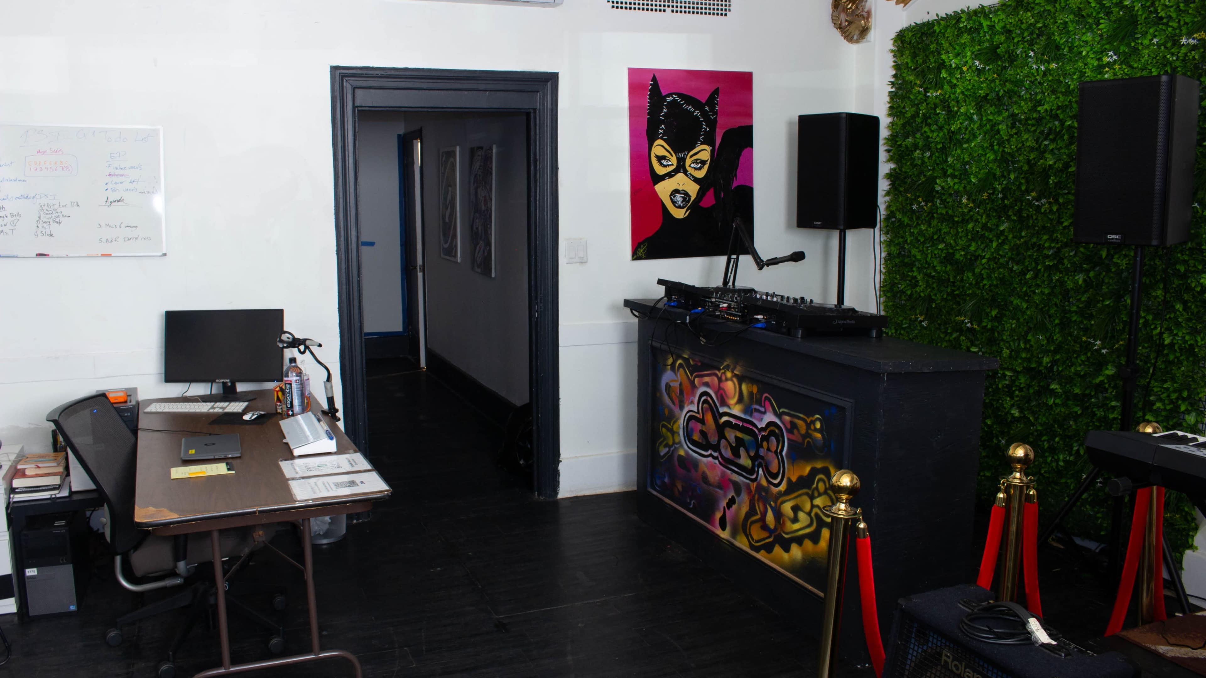 The image shows a room with a DJ setup featuring a black countertop, a colorful mural on the wall, an office desk, and a doorway leading to another space.