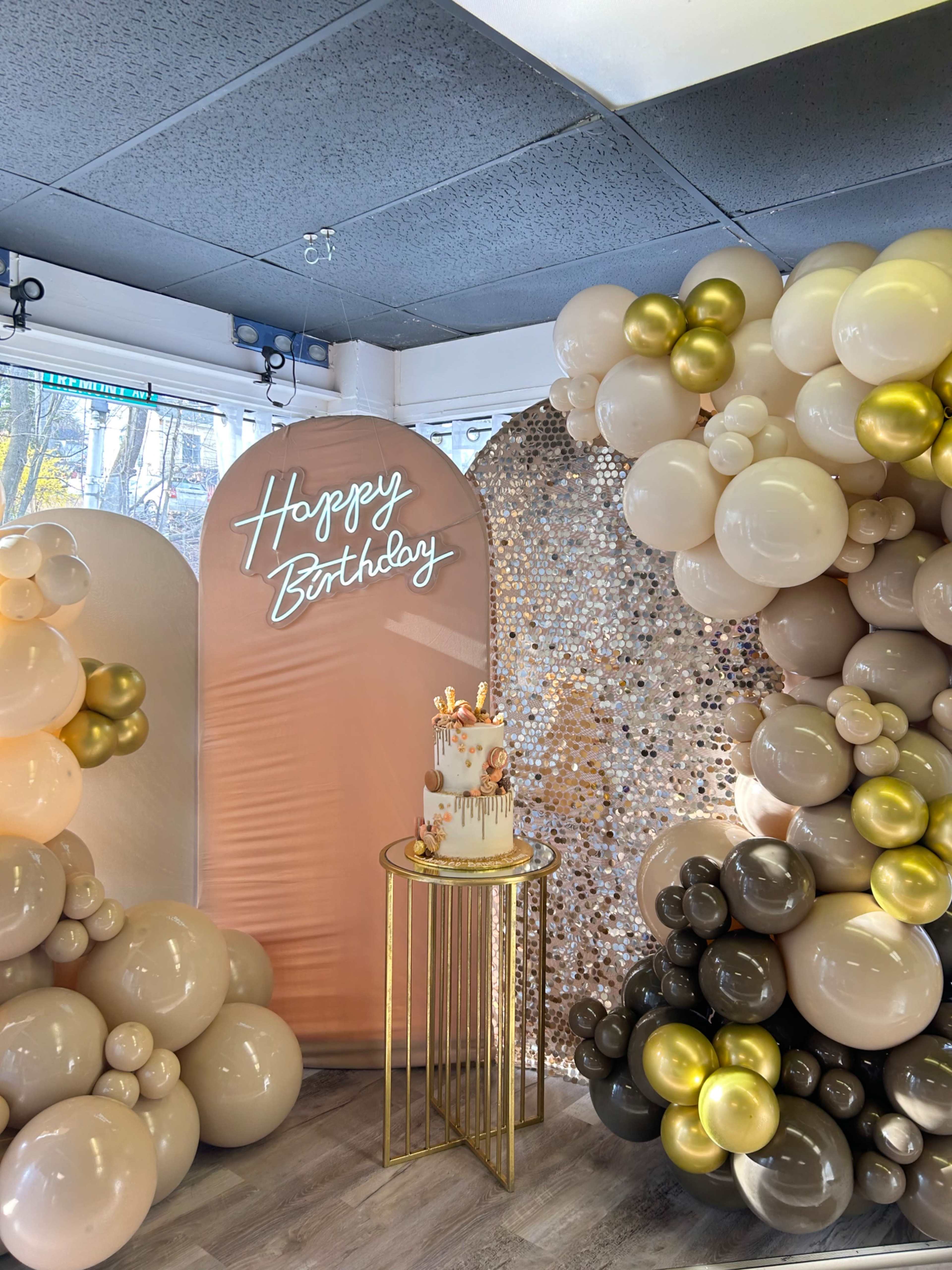 A birthday celebration display with a cake on a gold table, surrounded by a backdrop of balloons in various colors and a neon "Happy Birthday" sign.
